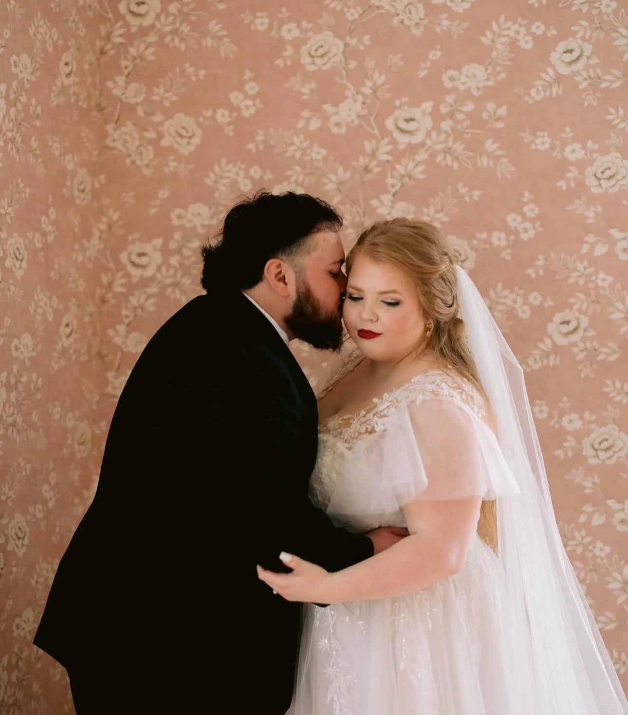 A bride and groom kissing on their wedding day, with the groom kissing the bride's temple, against a floral wallpaper background.