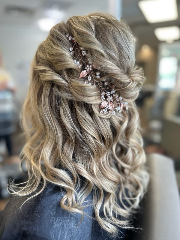 Blonde woman with curled hair styled in an updo adorned with a pink and crystal floral hair accessory, sitting indoors.