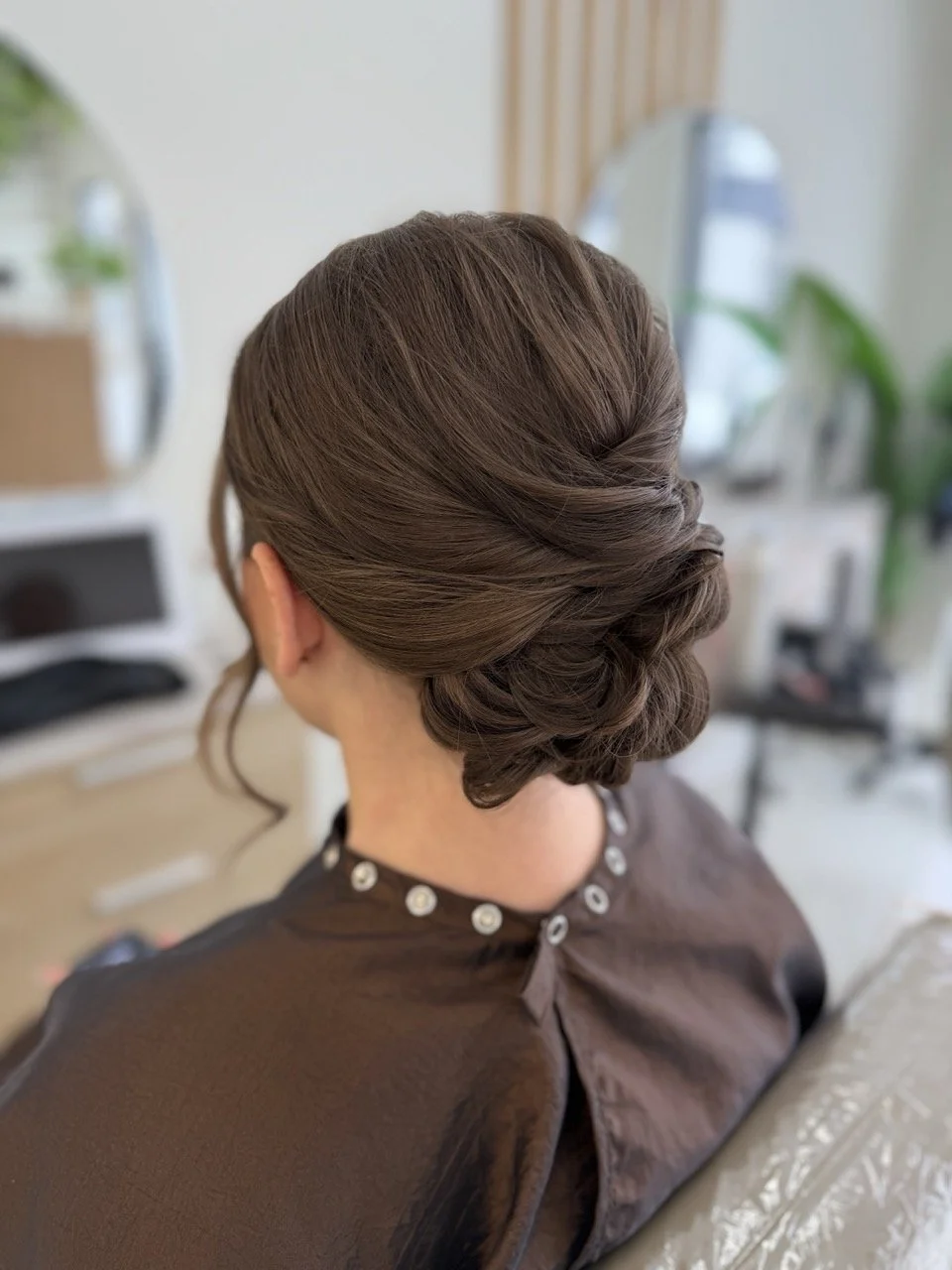 Woman with elegant brunette hairstyle sitting in a salon with mirrors and plants in the background.