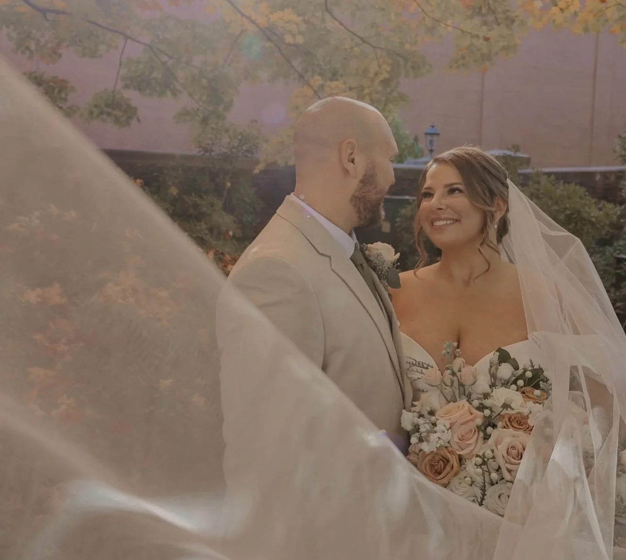 A bride and groom looking at each other, with the bride smiling and holding a bouquet of flowers, outdoors during their wedding.
