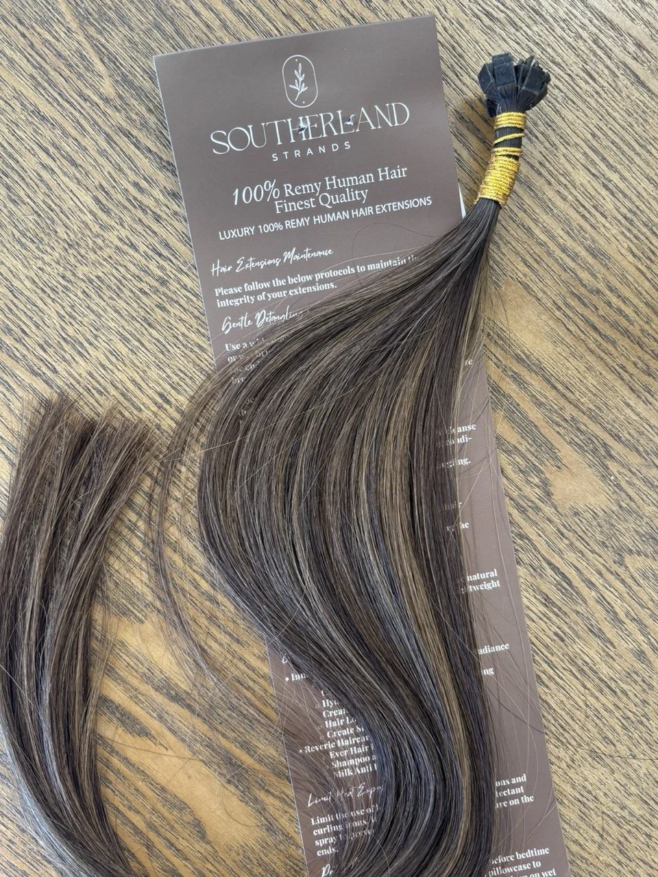 A bundle of Remy human hair extensions in shades of brown, secured with yellow and black bands, placed on a brown-colored informational card with the logo and text about hair care, on a wooden surface.