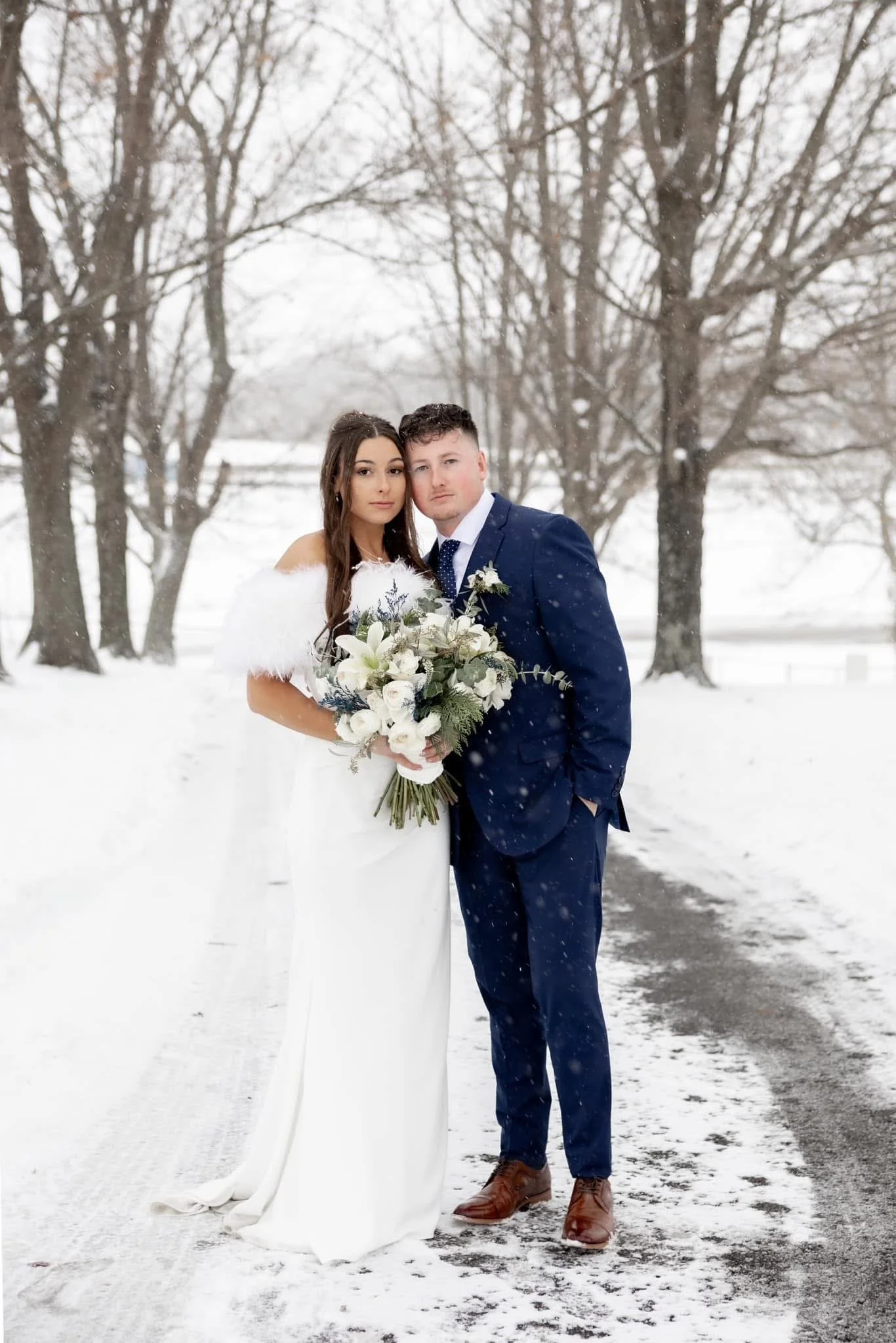 A bride and groom stand together outdoors in a snowy landscape, with bare trees in the background. The bride wears a white gown and holds a bouquet of white flowers, and the groom wears a navy blue suit with brown shoes.