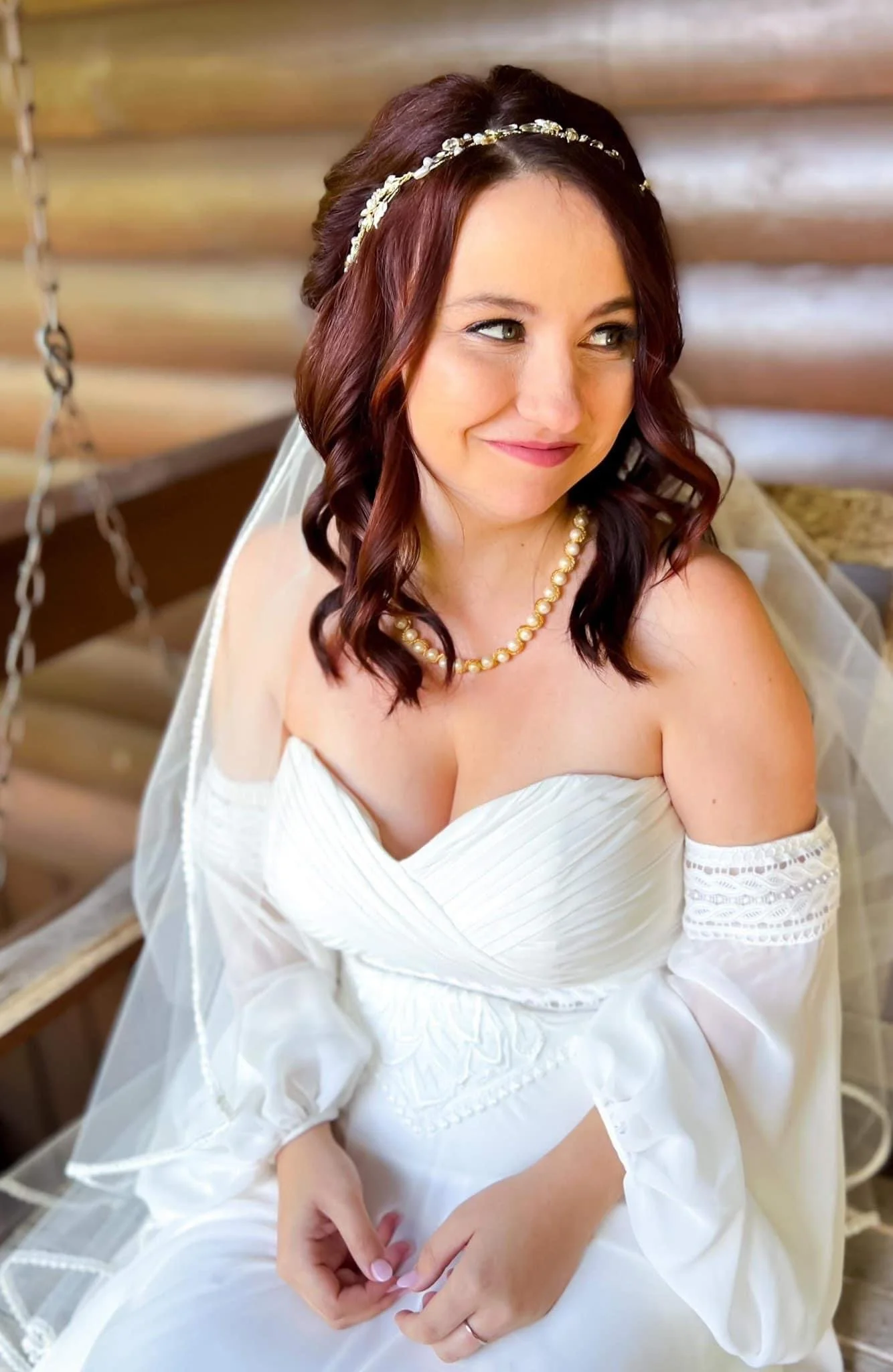 A bride with shoulder-length wavy brown hair wearing a white wedding dress, pearl necklace, veil, and tiara, sitting on a wooden bench.