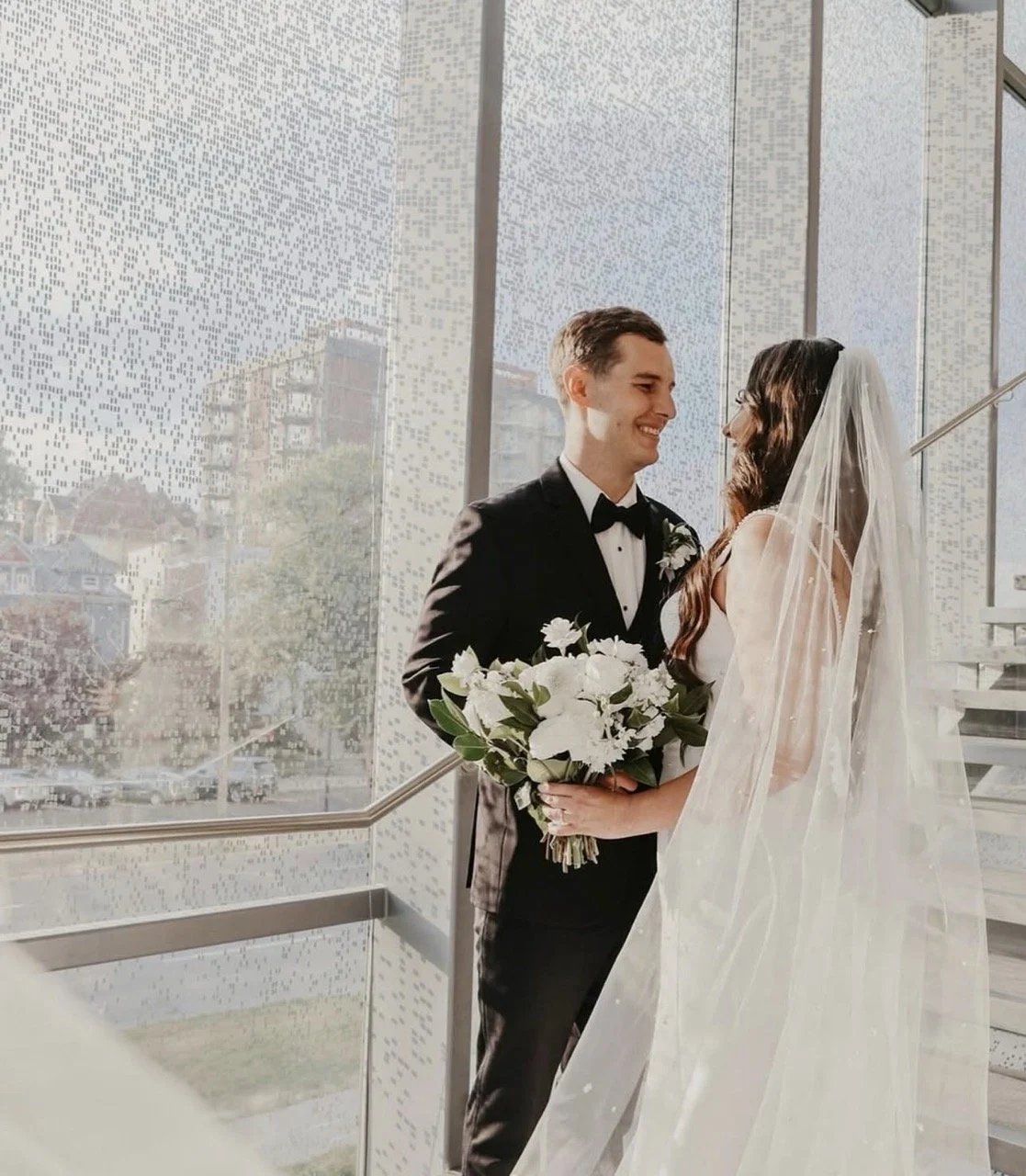 A bride and groom standing close indoors by a large window, smiling at each other with the groom holding a bouquet of white flowers, during their wedding.