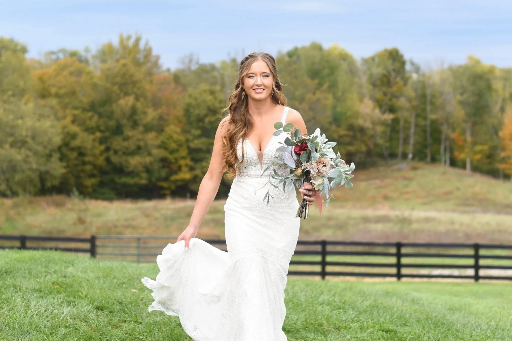 A bride wearing a white wedding dress with lace details, holding a bouquet of flowers, standing on a grassy field with trees and a fence in the background.