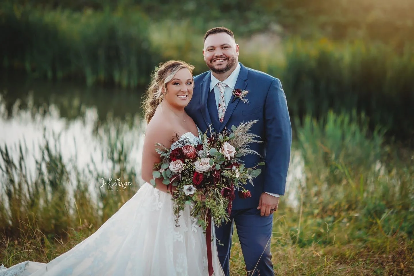 A smiling bride and groom standing outdoors near a lake, with the bride holding a large bouquet of flowers and greenery, both dressed in wedding attire.