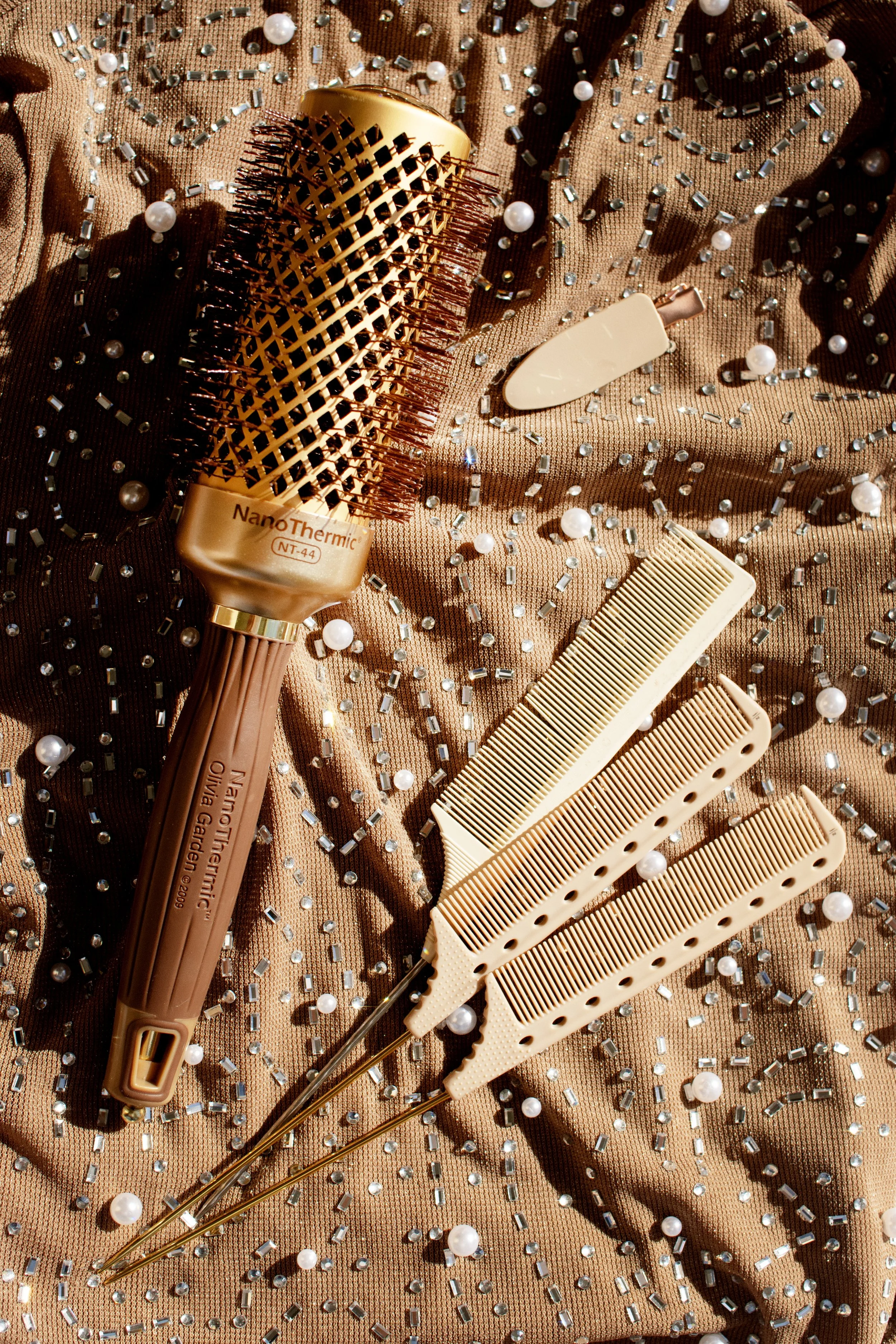 Hair styling tools including a round vented brush labeled NanoThermic NT-44, two beige hair clips, a beige hair comb, and a beige hair sectioning pick, all placed on a shiny, brown, sequined fabric background.