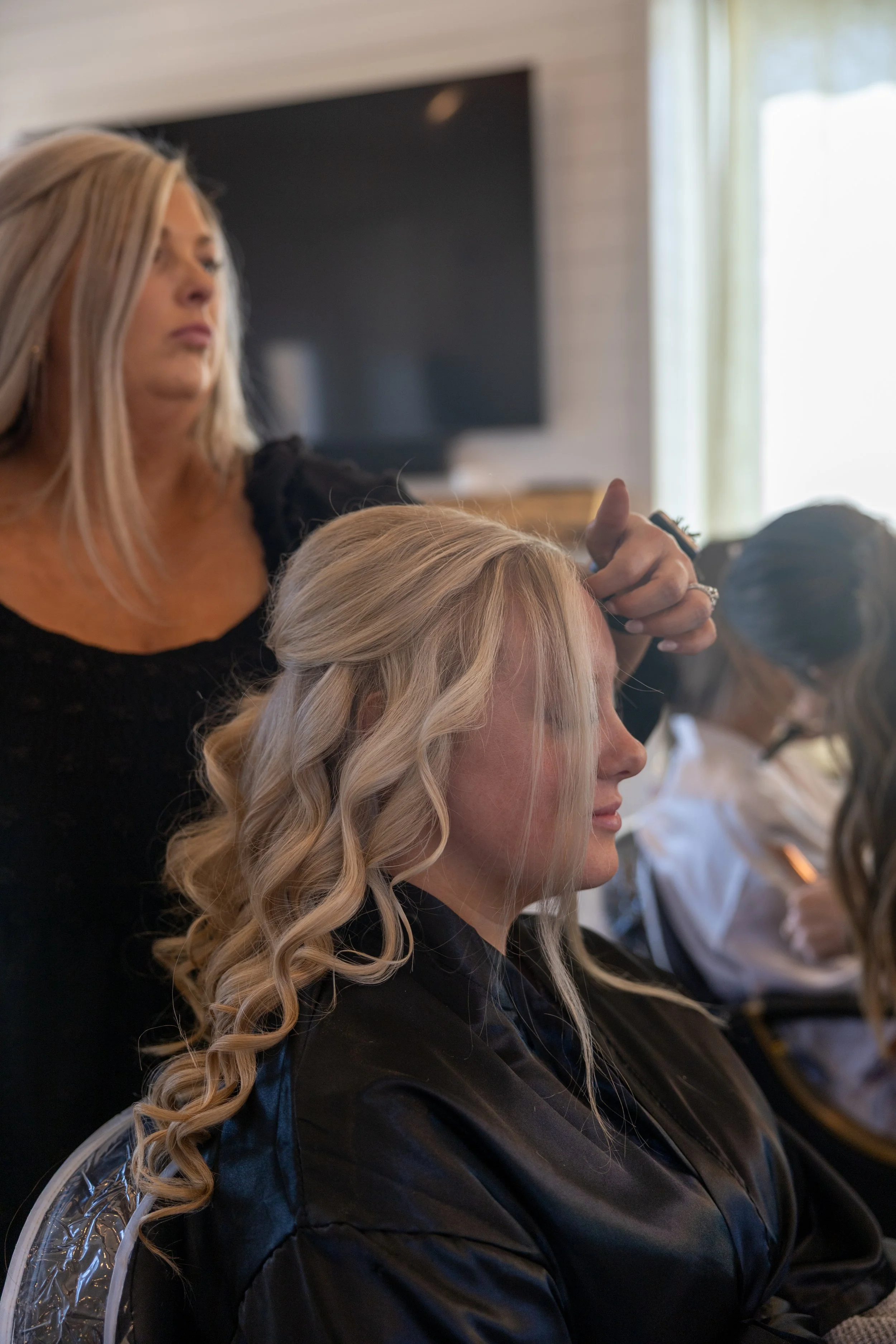 A woman with long blonde curly hair getting her hair styled by a hairstylist in a salon.