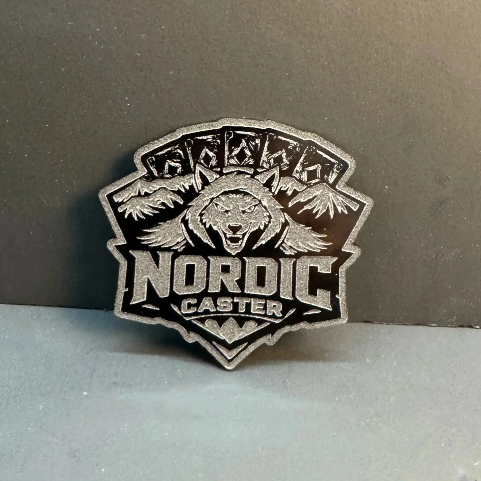 Insigne gravé pour une équipe de carte en forme de bouclier avec un loup et des montagnes, portant le texte « Nordest Caster »