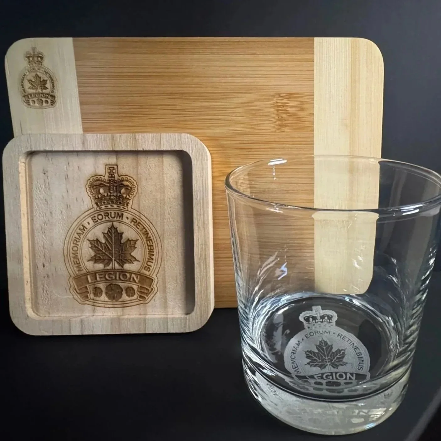 Ensemble cadeau. Verres et sous-verre et planche a fromage en bois avec inscriptions et monogrammes gravés, portant un logo de feuille d'érable, sur une surface noire.