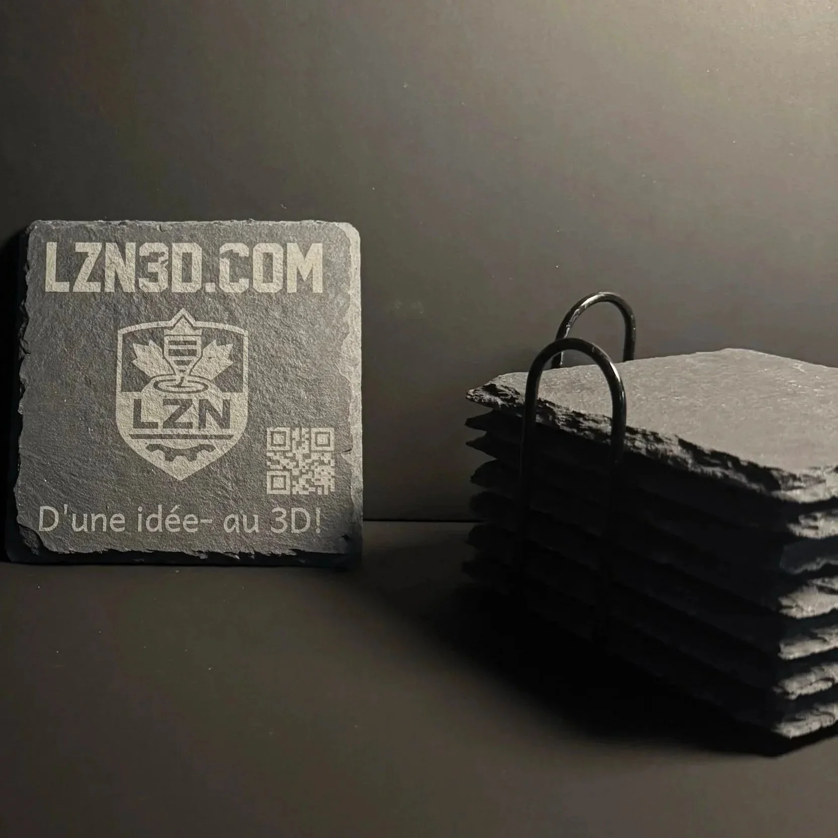 Ensemble de sous-verre en pierre avec l'inscription "LZN30.COM" un logo de feuille d'érable accompagnée du texte "D'une idée - au 3D!" gravé devant un mur gris sombre.