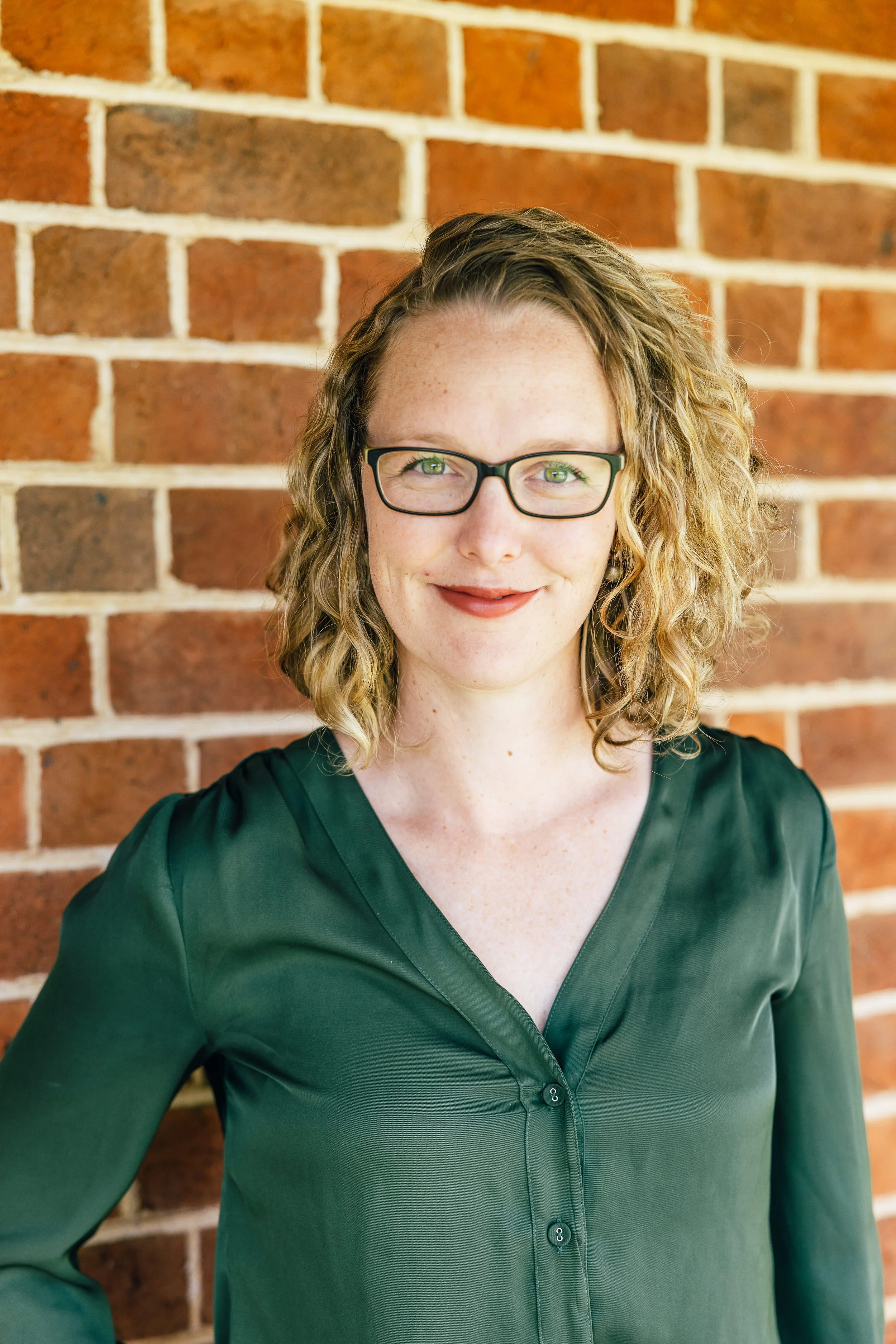 A woman with curly blonde hair, glasses, and a green blouse standing in front of a brick wall.