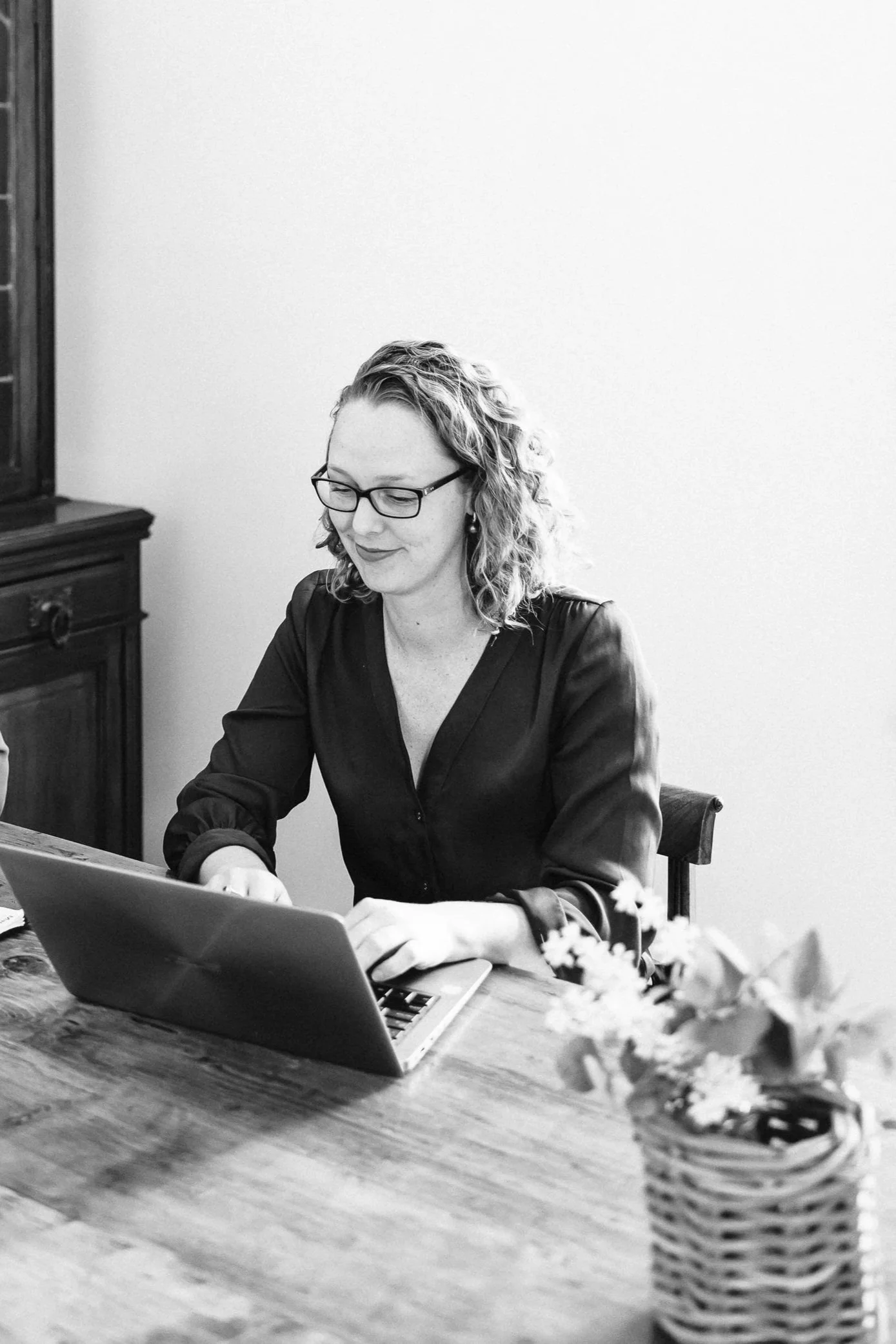 A woman with curly hair and glasses working on a laptop at a wooden table, with a vase of flowers nearby in a well-lit room.