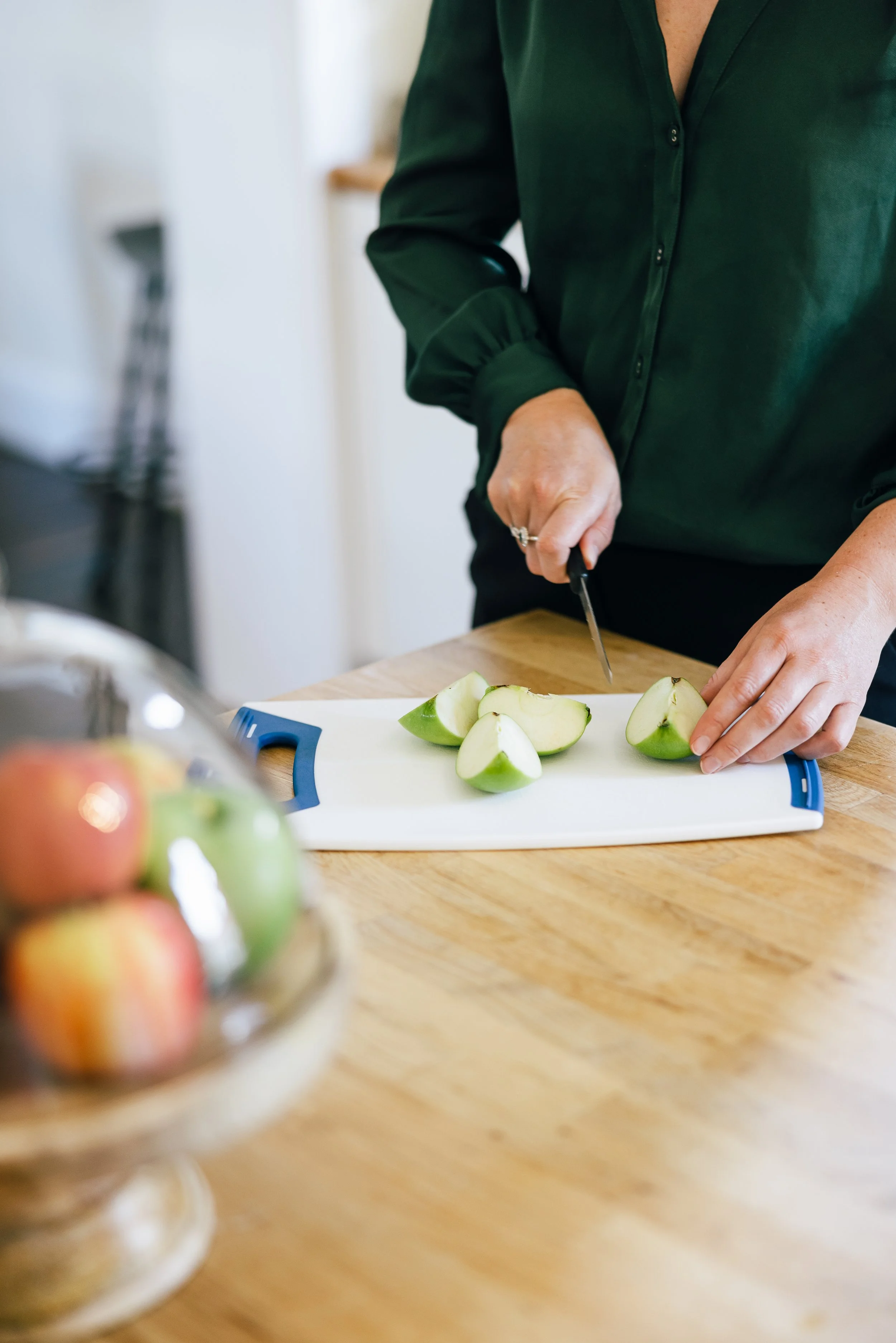 Person in a green shirt slicing a green apple on a cutting board in a kitchen.