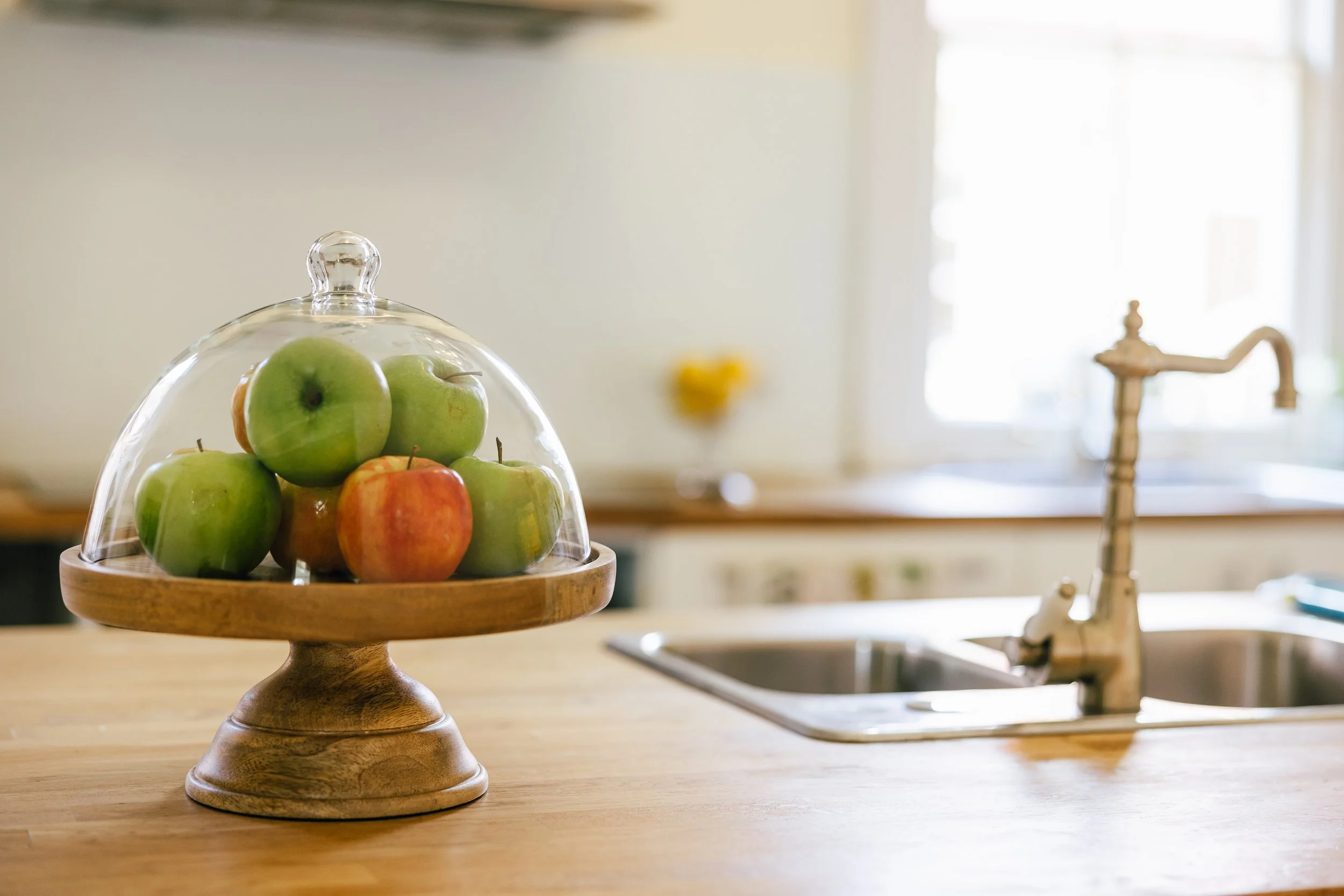A glass dome covering green and red apples on a wooden cake stand in a bright kitchen with a stainless steel sink and a brass faucet.