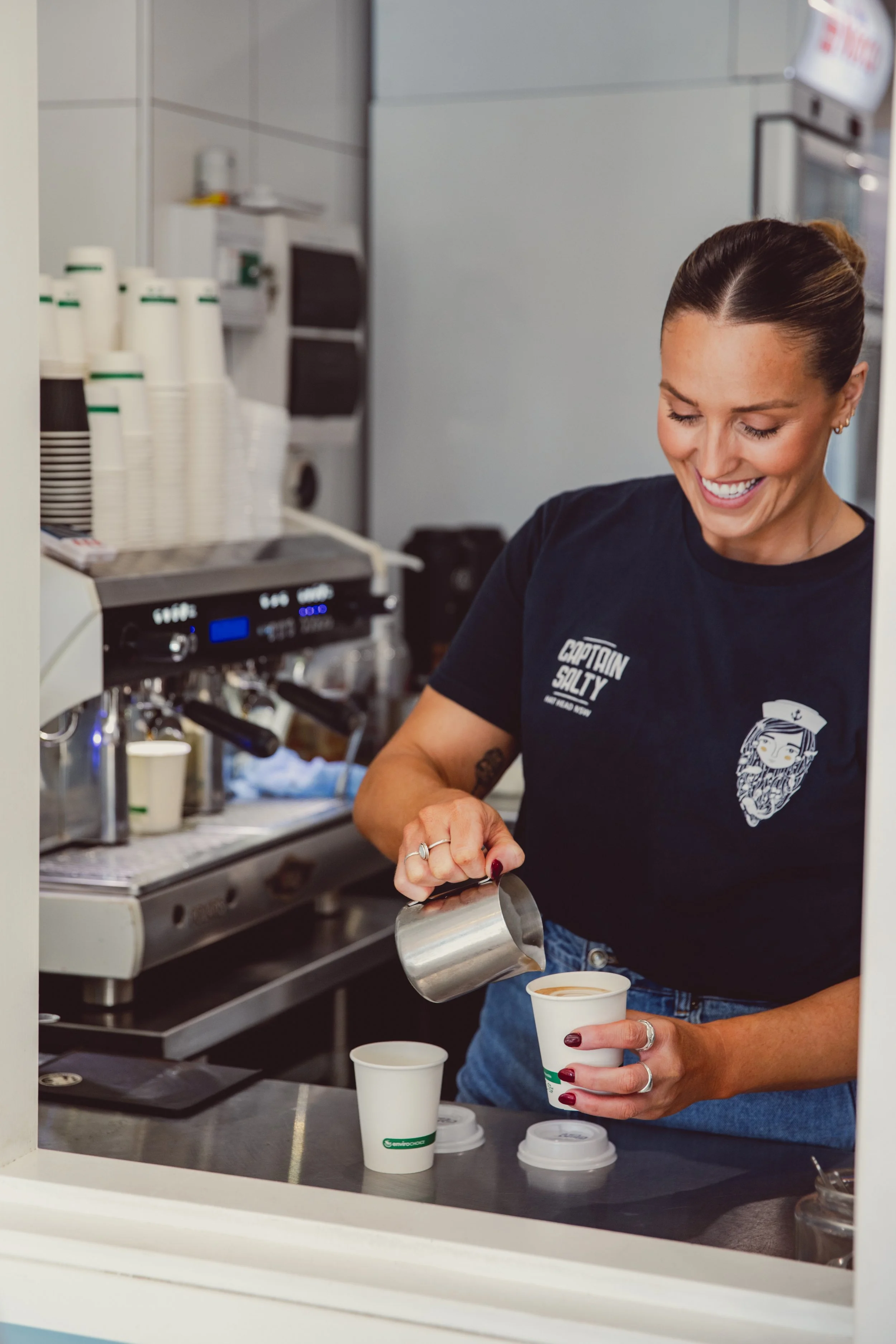 A smiling woman in a black t-shirt pours coffee from a metal pitcher into a paper cup at a coffee stand.