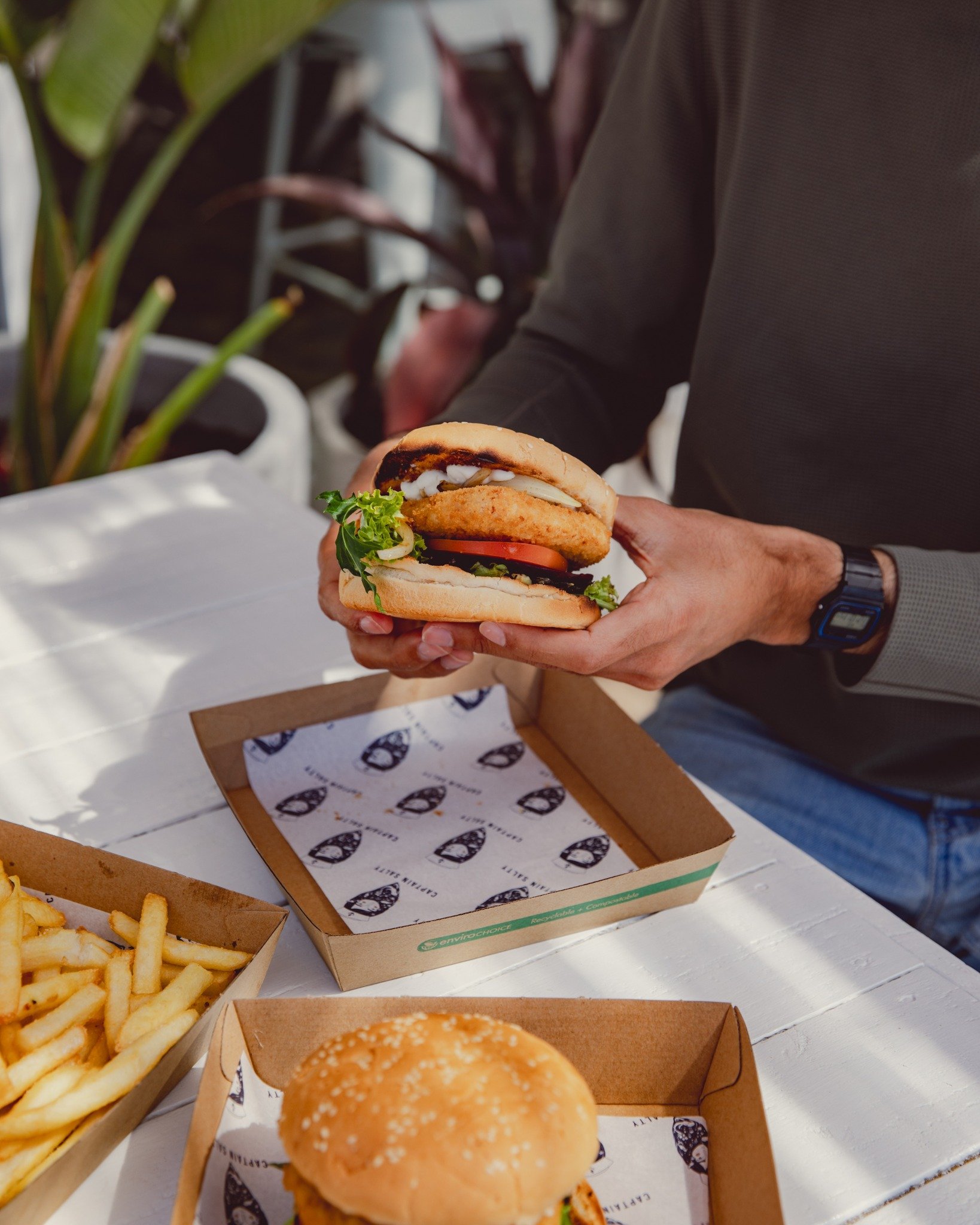 Person holding a burger with lettuce, tomato, cheese, and fried chicken patty, with fries and a burger on a white table, greenery in the background.