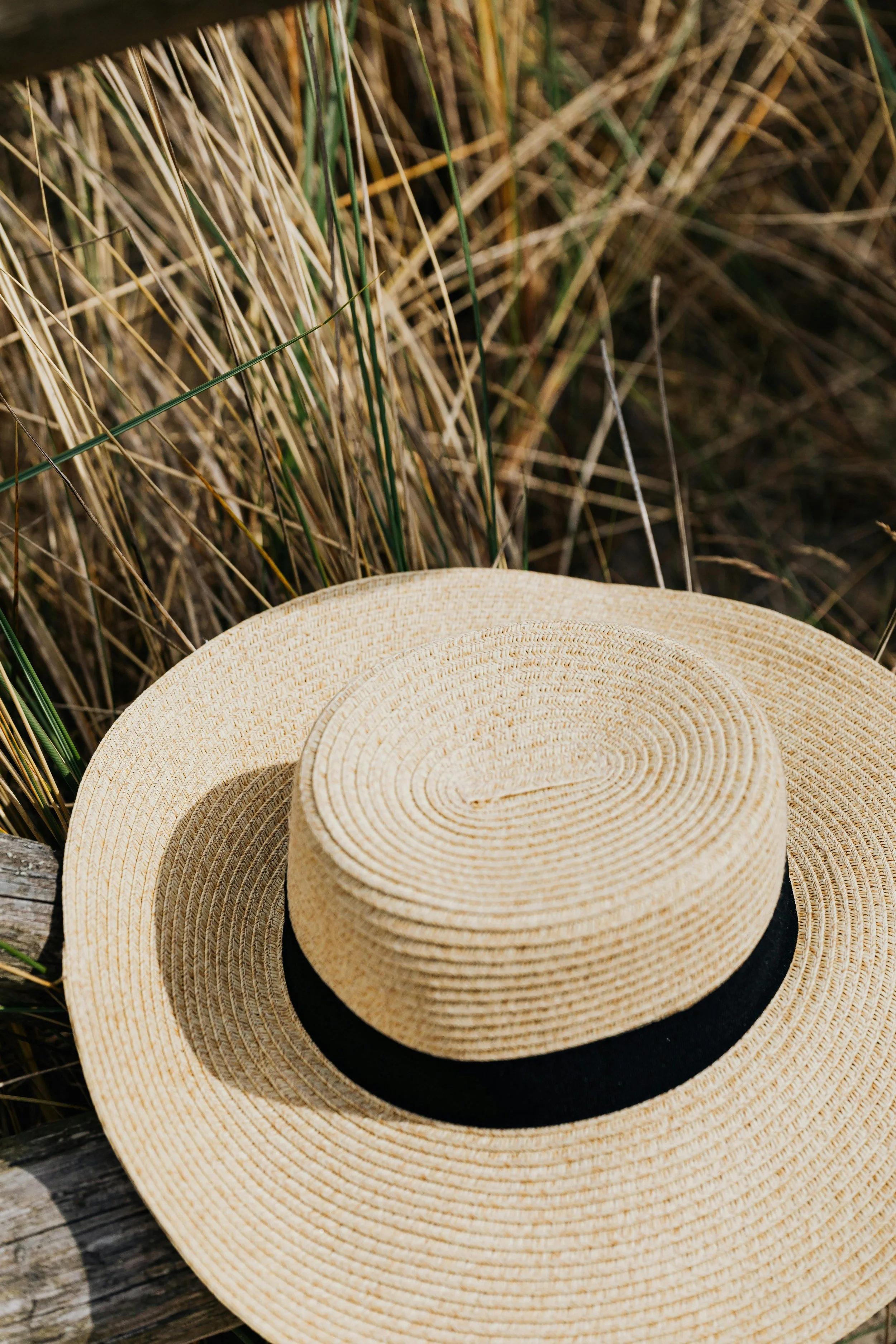 A straw sun hat with a black band resting on a weathered wooden surface in front of tall grassy plants.