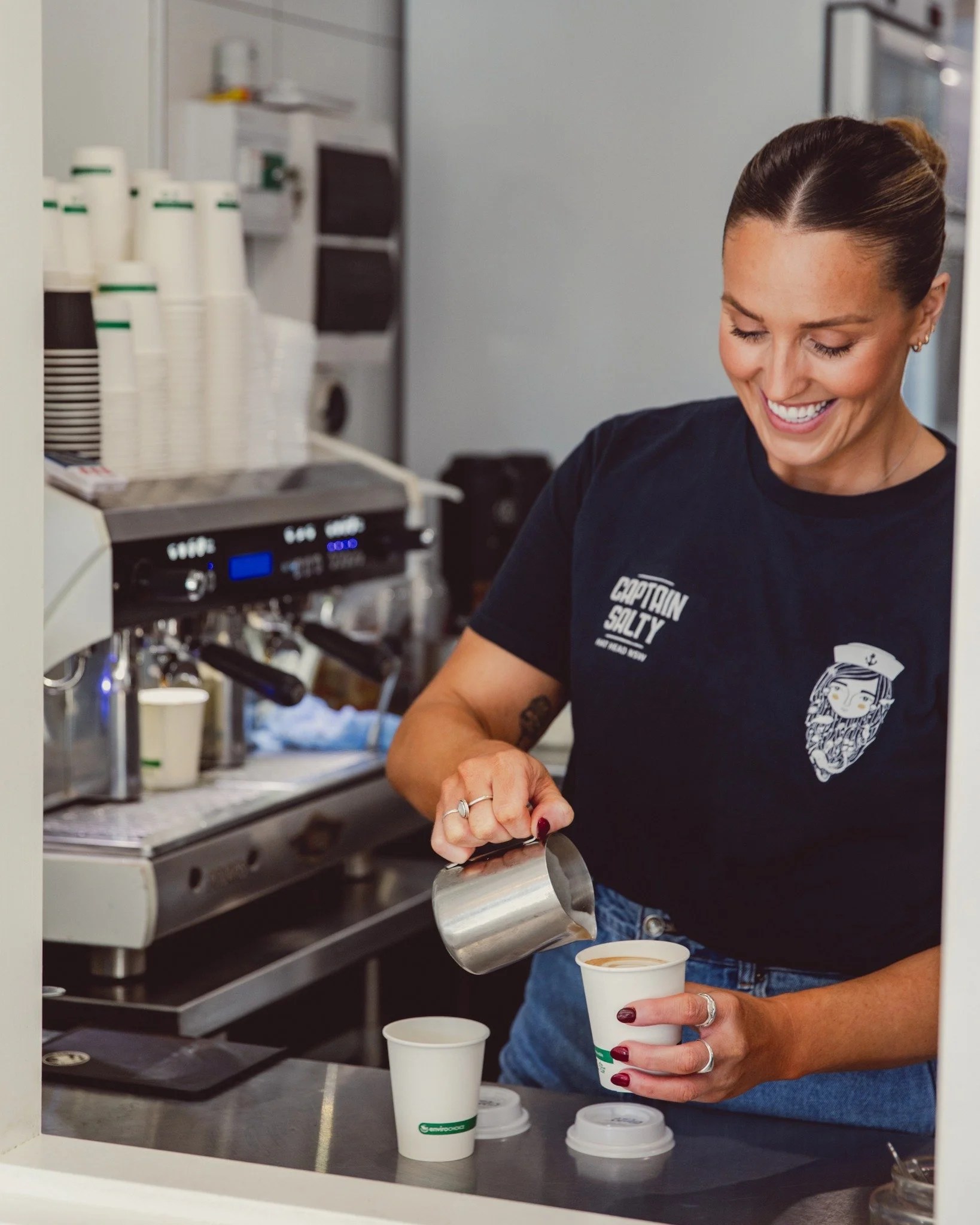 A woman smiling as she pours coffee into a paper cup at a coffee shop counter.