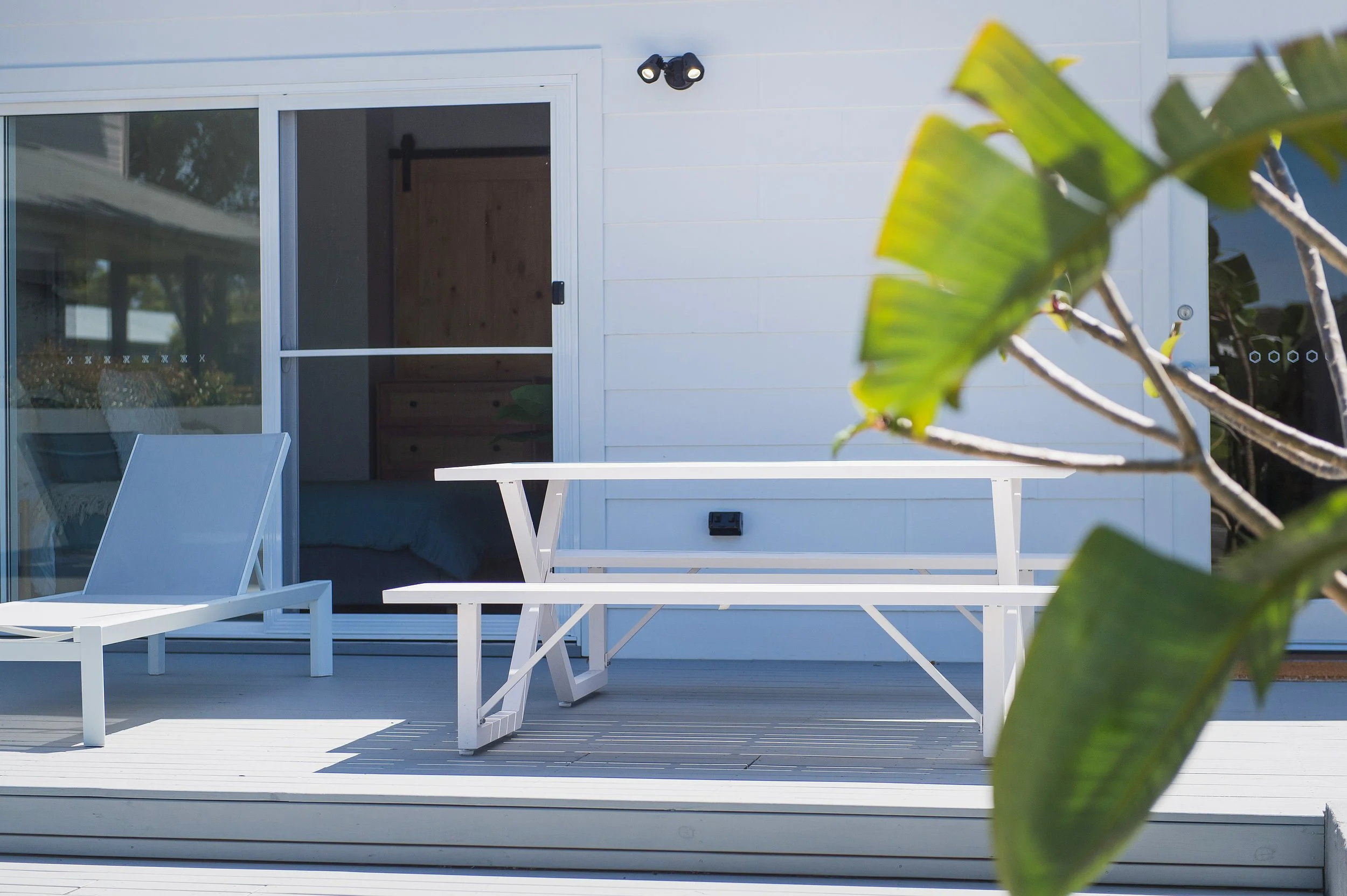 White outdoor patio furniture with a lounge chair and a table in front of a sliding glass door, with a plant in the foreground and a white house exterior.