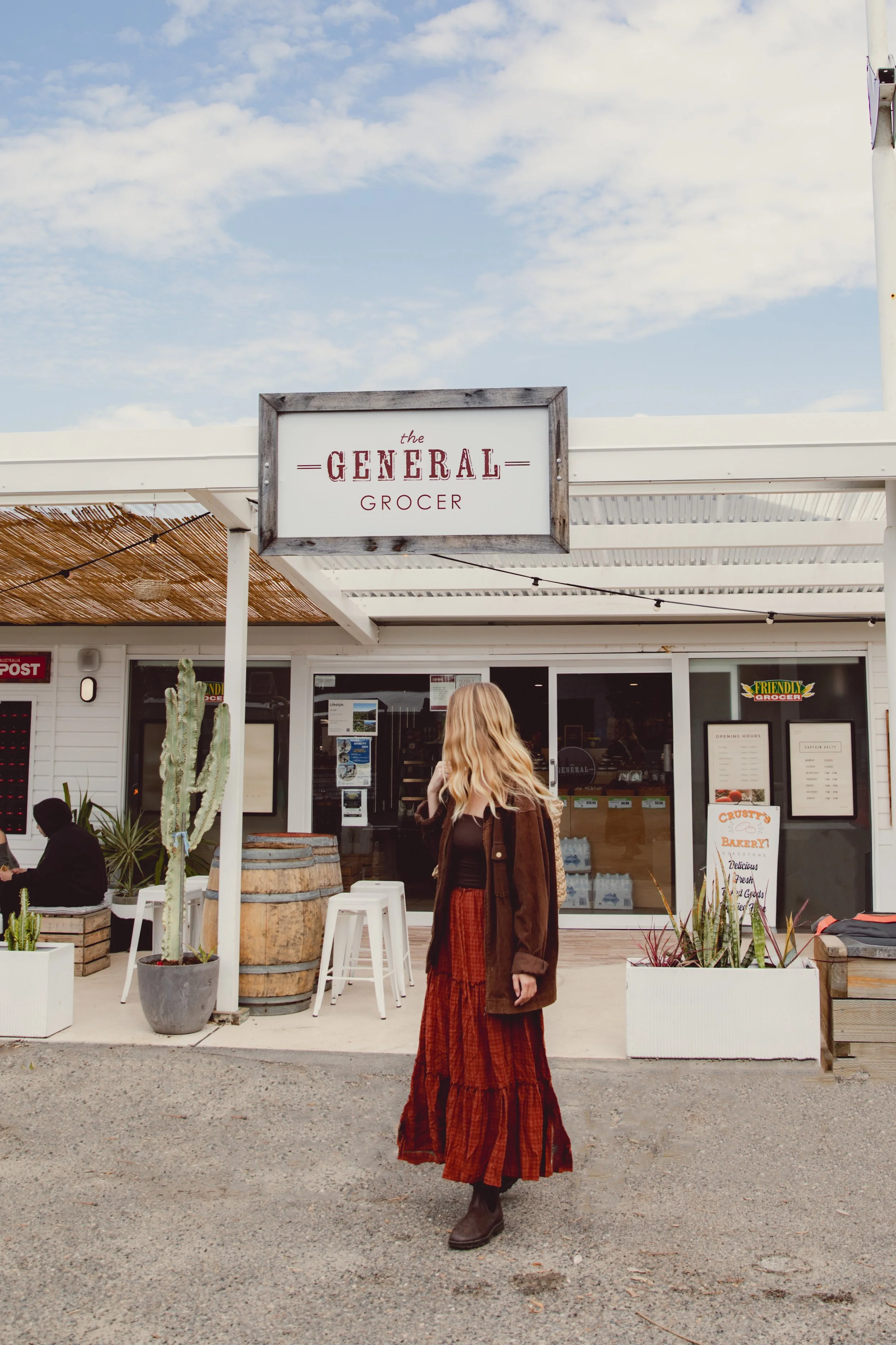 A woman with long blonde hair wearing a brown coat, black top, and a long rust-colored skirt standing outside a store called 'The General Grocer' with a cactus plant in a pot nearby.