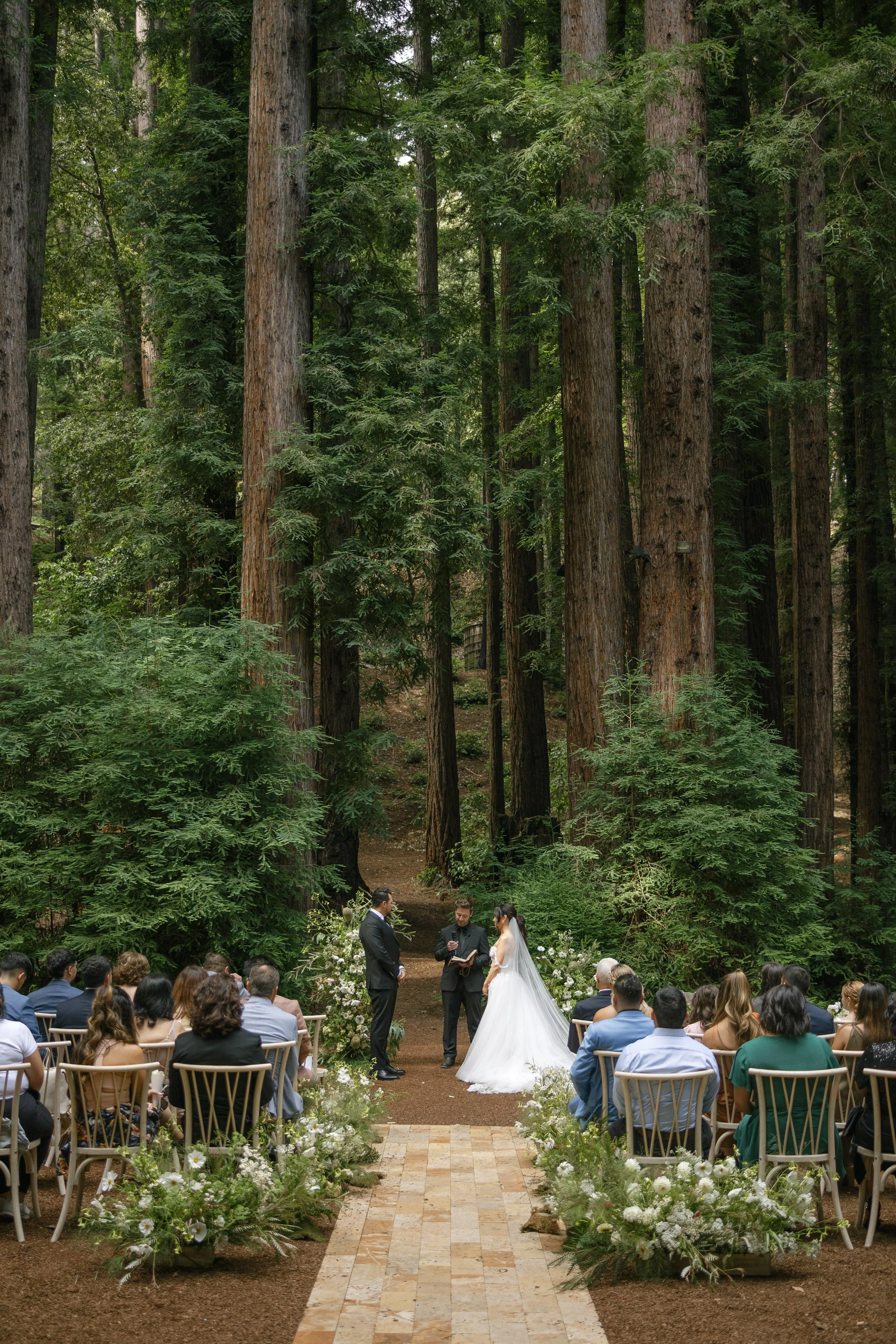 A wedding ceremony taking place outdoors in a forest, with the bride and groom standing before an officiant. Guests are seated in rows on either side, surrounded by tall trees and lush greenery. The aisle is decorated with white flowers and greenery.