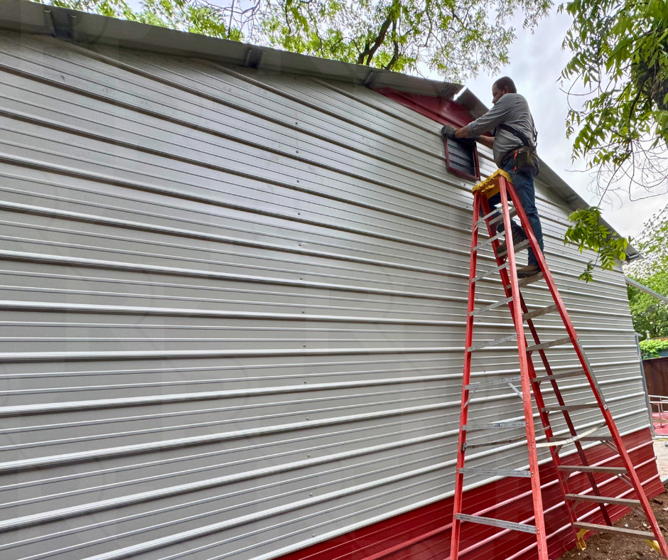 A man on a red ladder installing a window on the side of a house with metal siding.