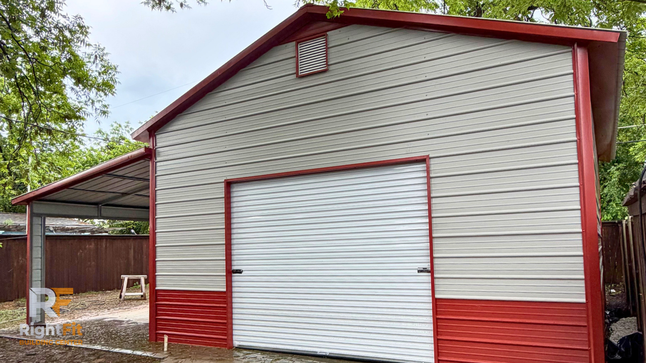 A metal garage with a white roll-up door, beige siding with red trim, and a small ventilation vent near the peak of the roof. There is a smaller covered area attached on the left side. Trees are visible in the background.