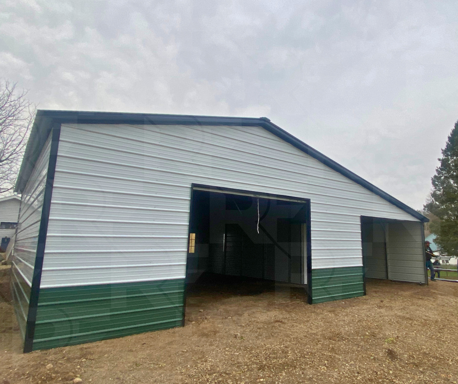 A green & white large metal shed with two open bays, one larger than the other, on a dirt ground with a cloudy sky overhead.