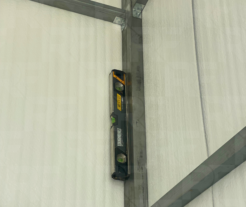 A black TrowelMate spirit level with yellow and green accents resting on a metal frame in front of a white wall.