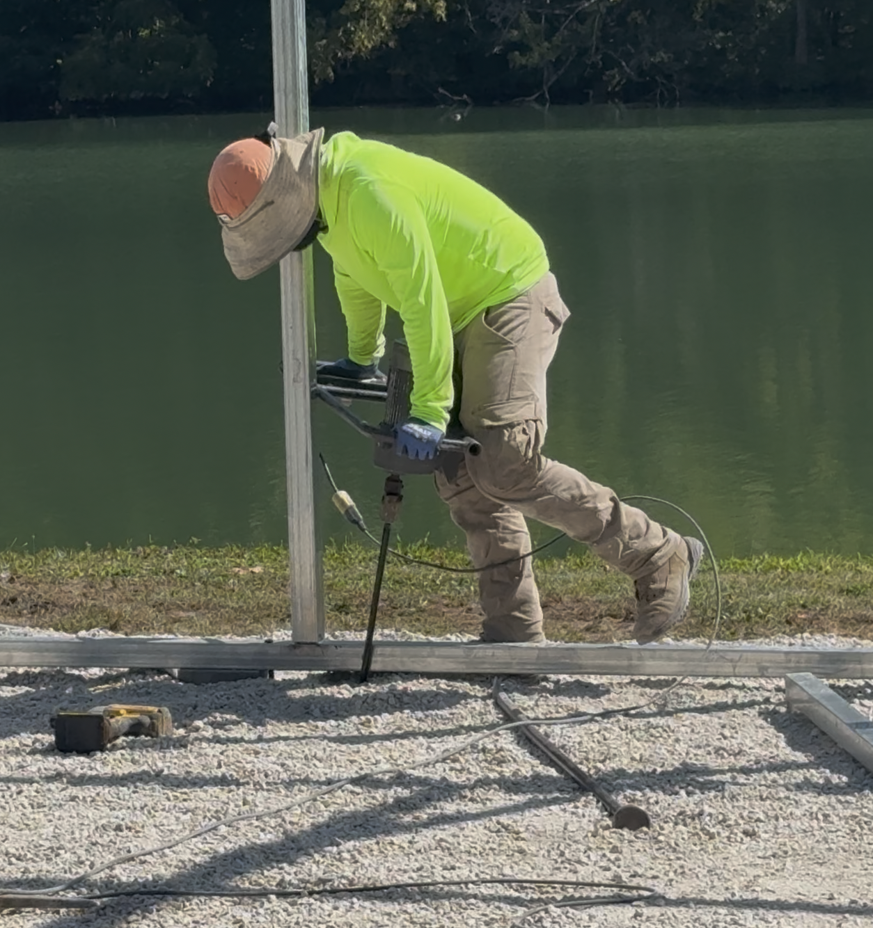 A worker in a bright yellow shirt, beige pants, and a wide-brimmed hat is installing or repairing a metal pole near a body of water. The worker is kneeling on gravel, using a power drill, with tools and wires on the ground.