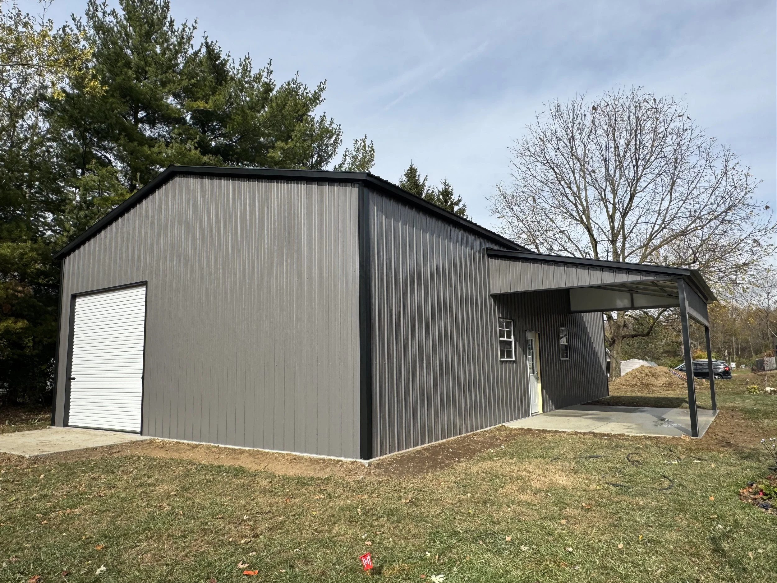 A gray metal building with a roll-up garage door and a side door, situated on a grassy area with trees in the background.