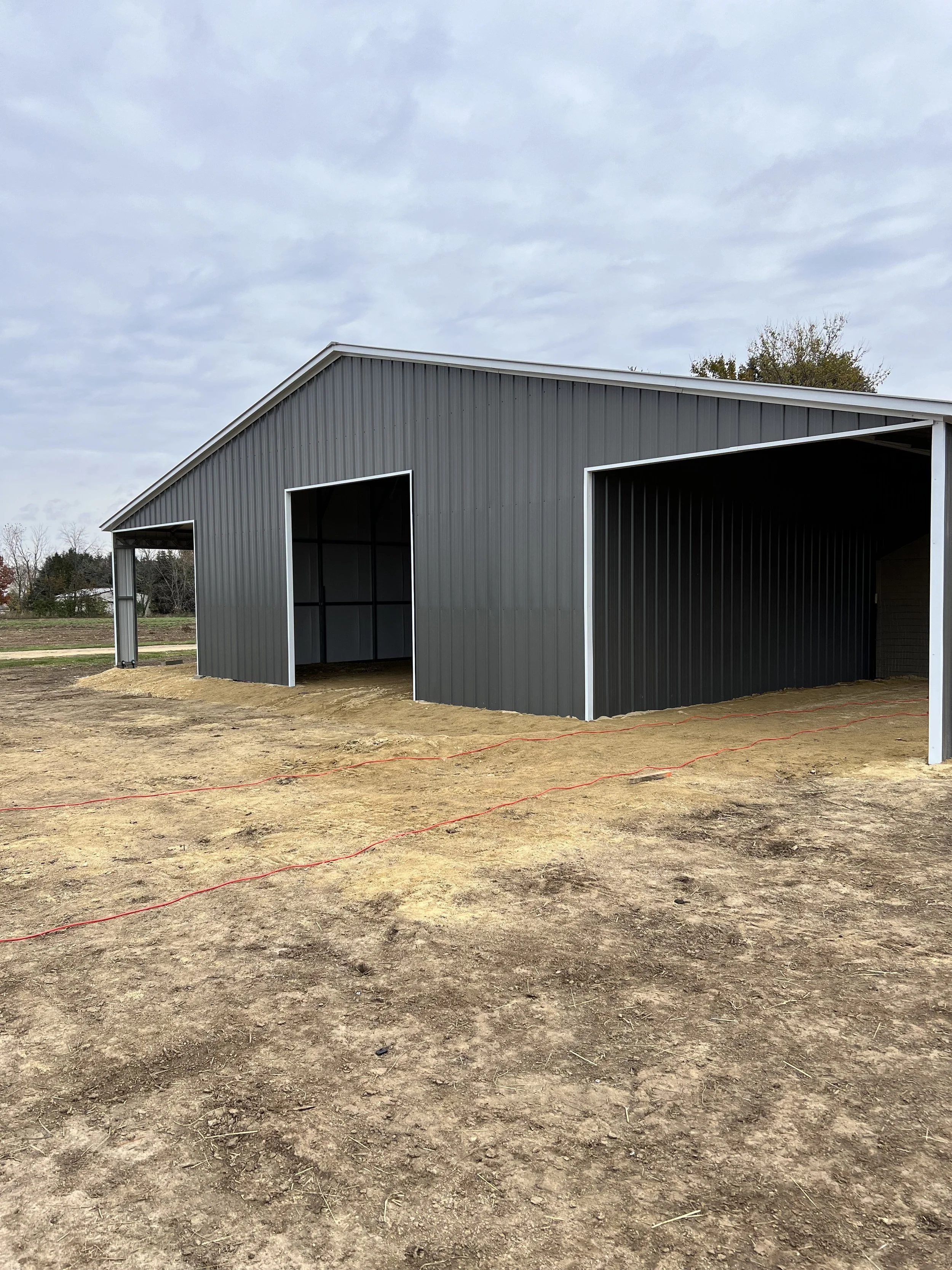 A metal building under construction with two large open garage doors, situated on a dirt lot with cordon strips on the ground, and a cloudy sky overhead.