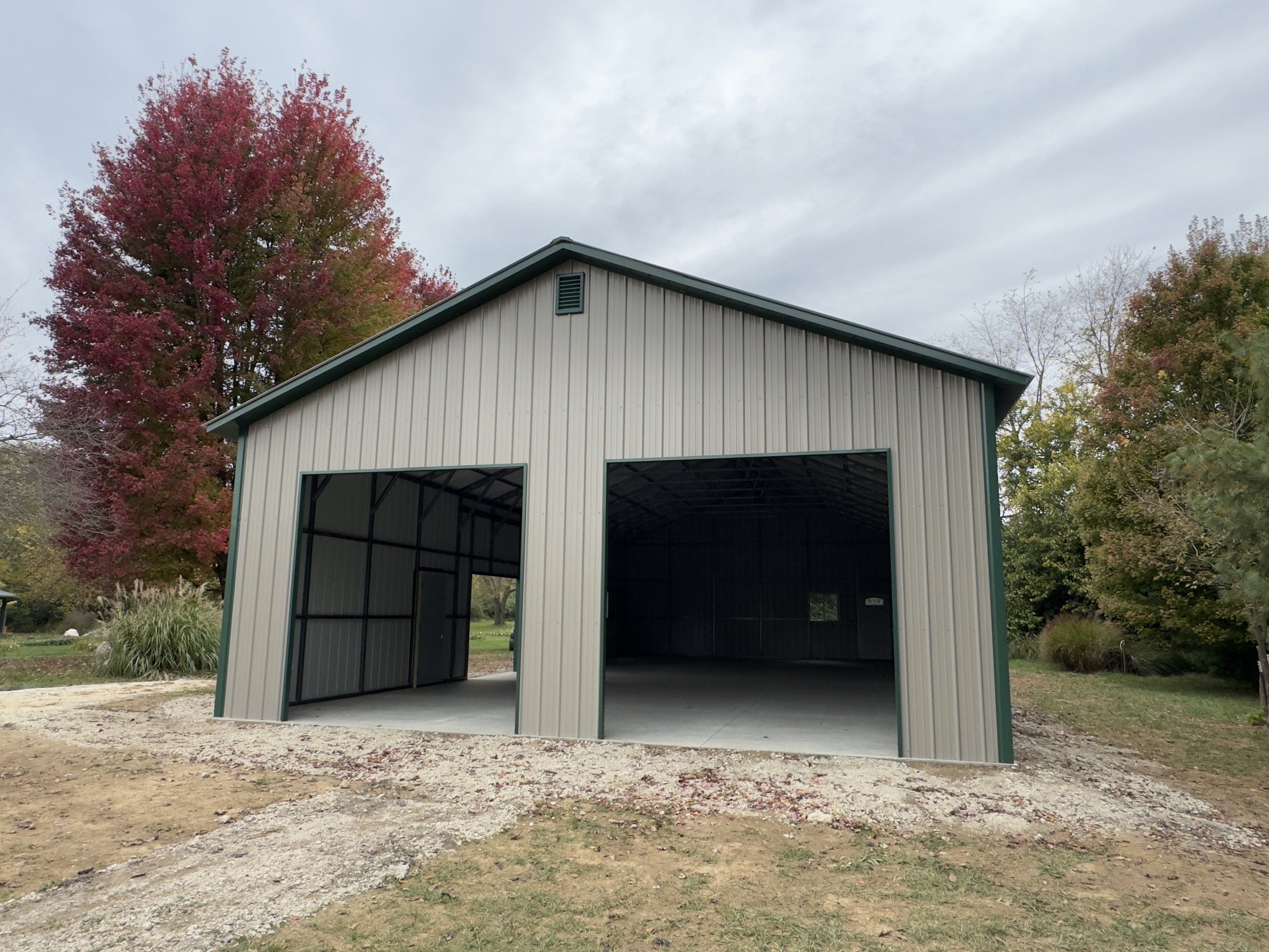 A large, empty metal framed garage or storage building with an open front, situated on a grassy area with trees in fall foliage in the background.