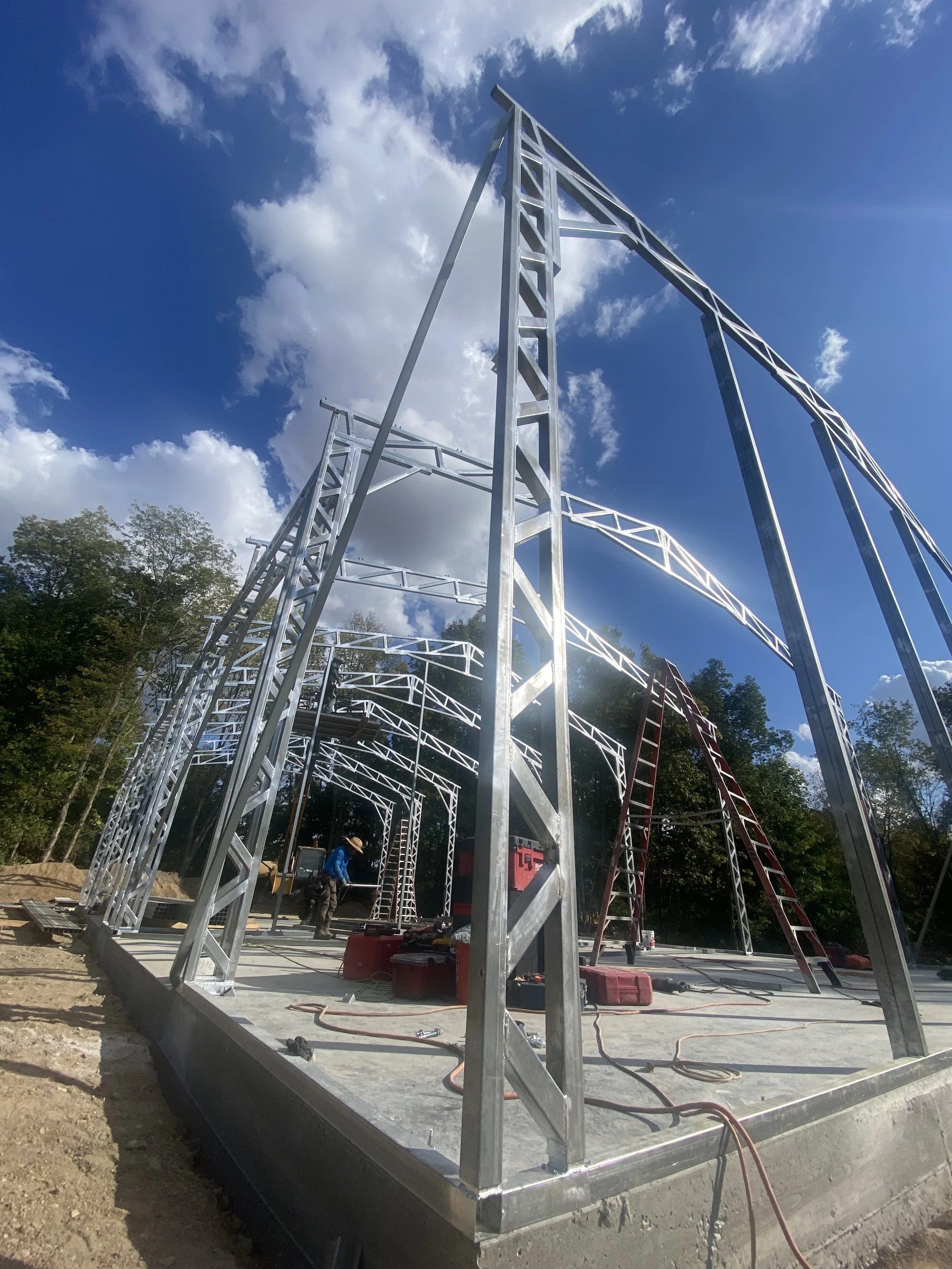 Construction site with metal framing structure, workers, ladders, and construction tools under a blue sky with clouds.