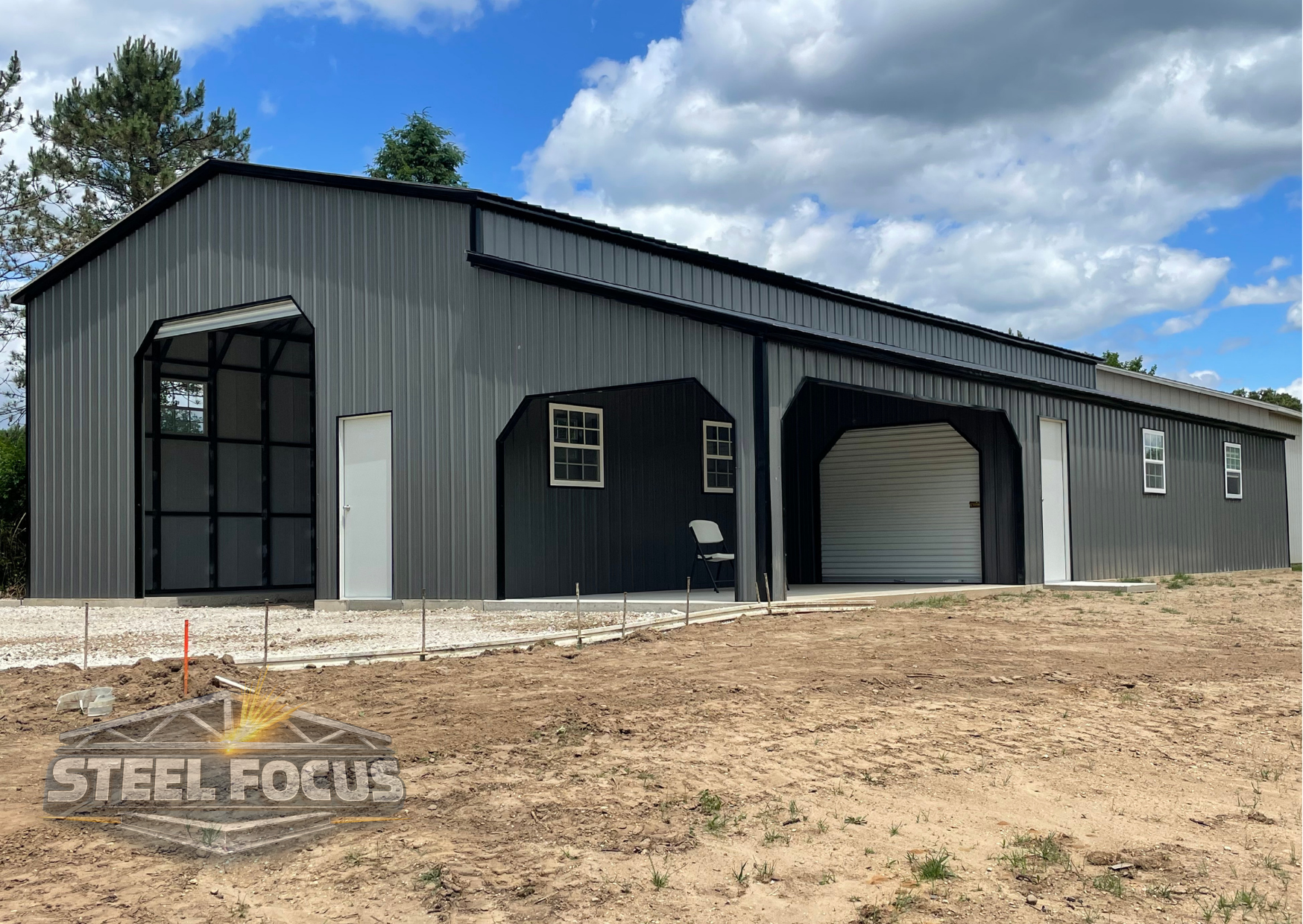 Large metal building with multiple doors and windows, on a dirt lot under a partly cloudy sky.
