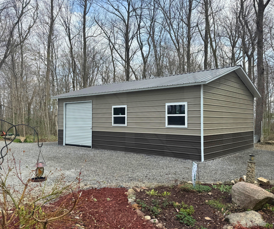 A metal storage shed with a cream and dark brown exterior, two windows, and a white roll-up door, situated on a gravel surface in a wooded area.