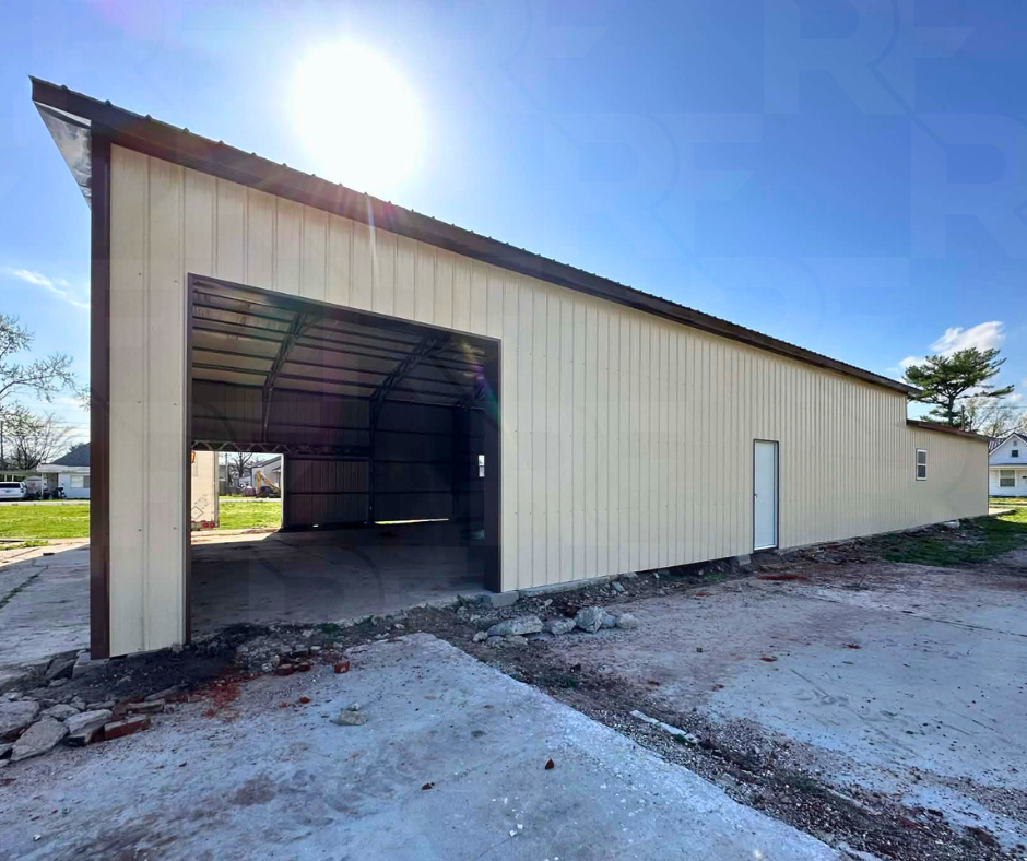 A large beige metal building with a large open garage door and a smaller door on the side, set on a construction site with dirt and rocks in front, under a sunny sky.