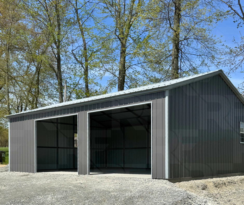 A dark gray vertical sided metal garage with two open garage bays, set against a background of tall trees and a clear blue sky.