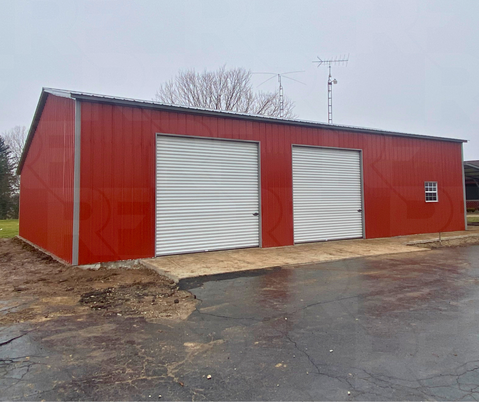 Red metal building with two white roll-up garage doors and a small window, set on a paved and gravel surface.