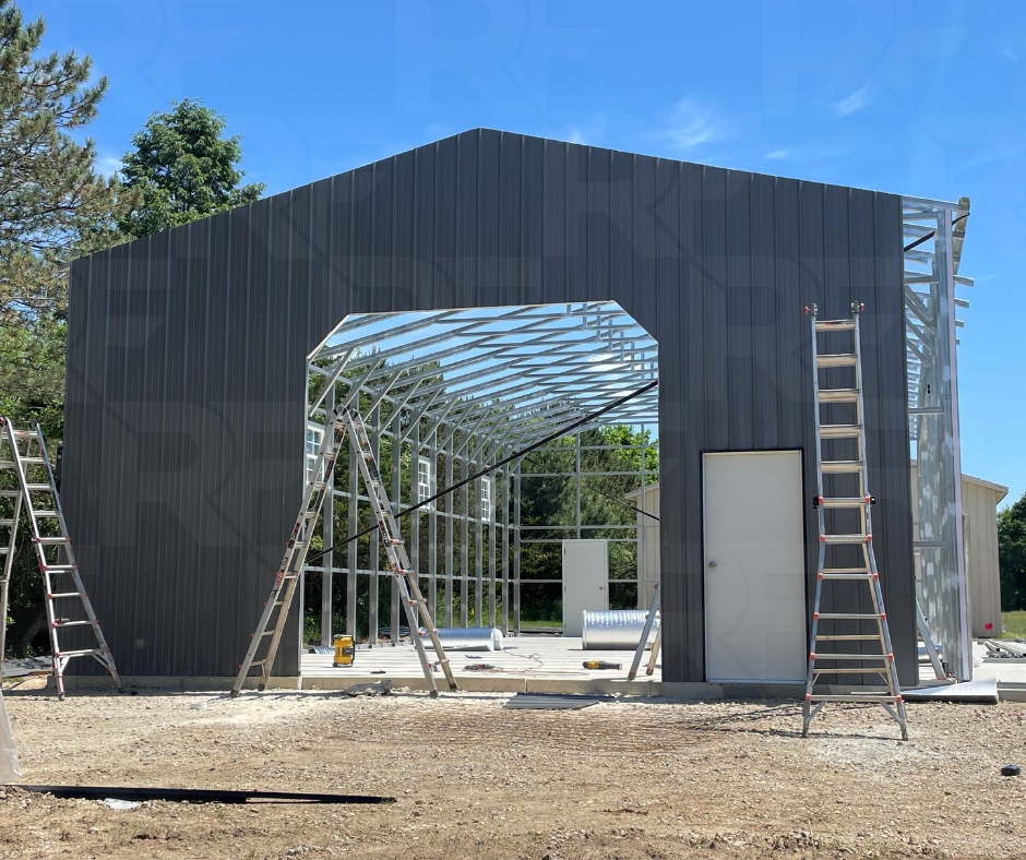 Steel building under construction with open framework, ladders leaning against it, and construction tools on the ground on a sunny day.