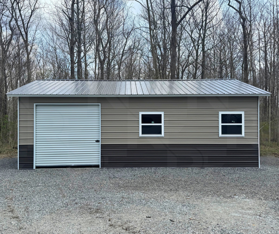 A small metal building with a gray roof, beige upper walls, and black lower walls, featuring a garage door and two windows, set against a background of leafless trees on a gravel lot.