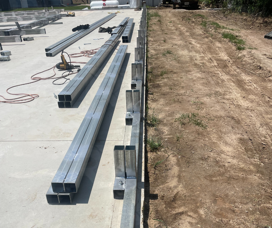 Construction site with metal framing materials, tools, and equipment on a concrete slab, next to a dirt pathway.