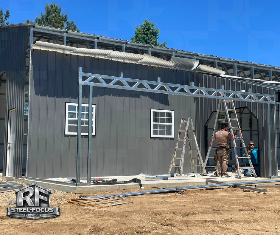 Construction workers installing a metal framework on a gray building with two small windows. There are ladders and construction tools, with a dirt ground in the foreground and trees in the background.
