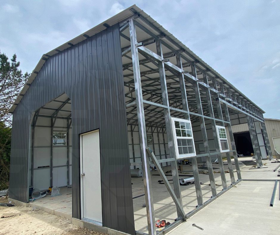 A metal building under construction, with a black metal exterior on one side and a metal frame on the other, two windows installed, and construction tools and materials on the ground.