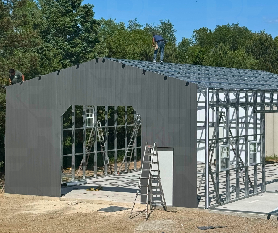 Construction workers building the frame and installing metal panels on a large metal building with multiple windows and a pitched roof, on a clear day surrounded by trees.