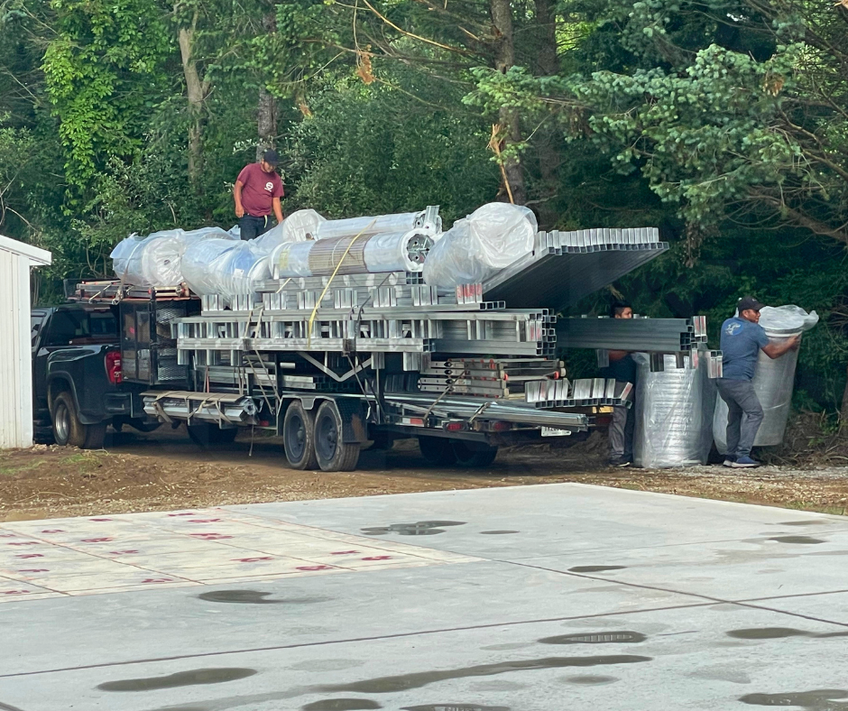 Workers unloading large rolls of insulation and metal framing materials from a truck near a wooded area.