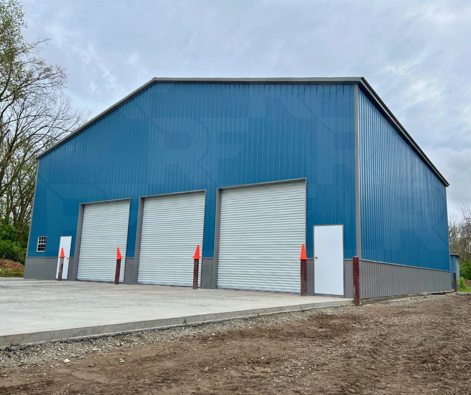 A blue 60x60x20 metal building with three large white garage doors, a side white door, and a small window, surrounded by a concrete driveway and some trees on the left.
