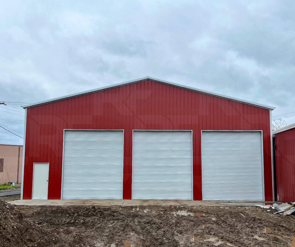 Red metal industrial building with three large white roll-up garage doors and a small side door, under a cloudy sky.