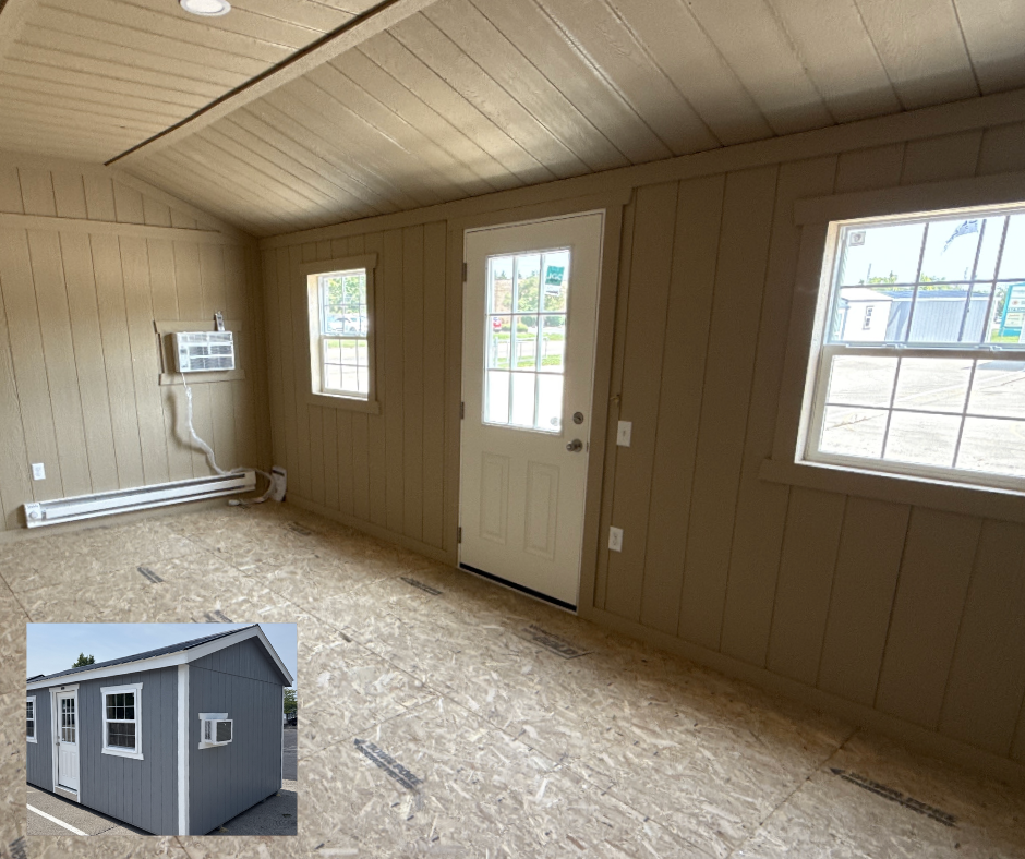 Interior of a small, unfinished tiny house with beige wooden panel walls, a white door with glass panes, three windows, and a wall-mounted air conditioning unit. The floor is unfinished plywood.